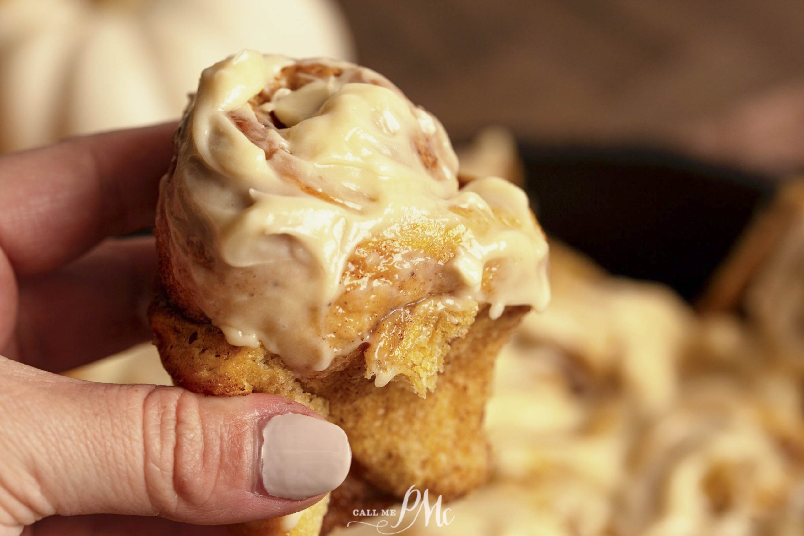 Close-up of a hand holding a cinnamon roll topped with cream cheese frosting, with more cinnamon rolls blurred in the background.