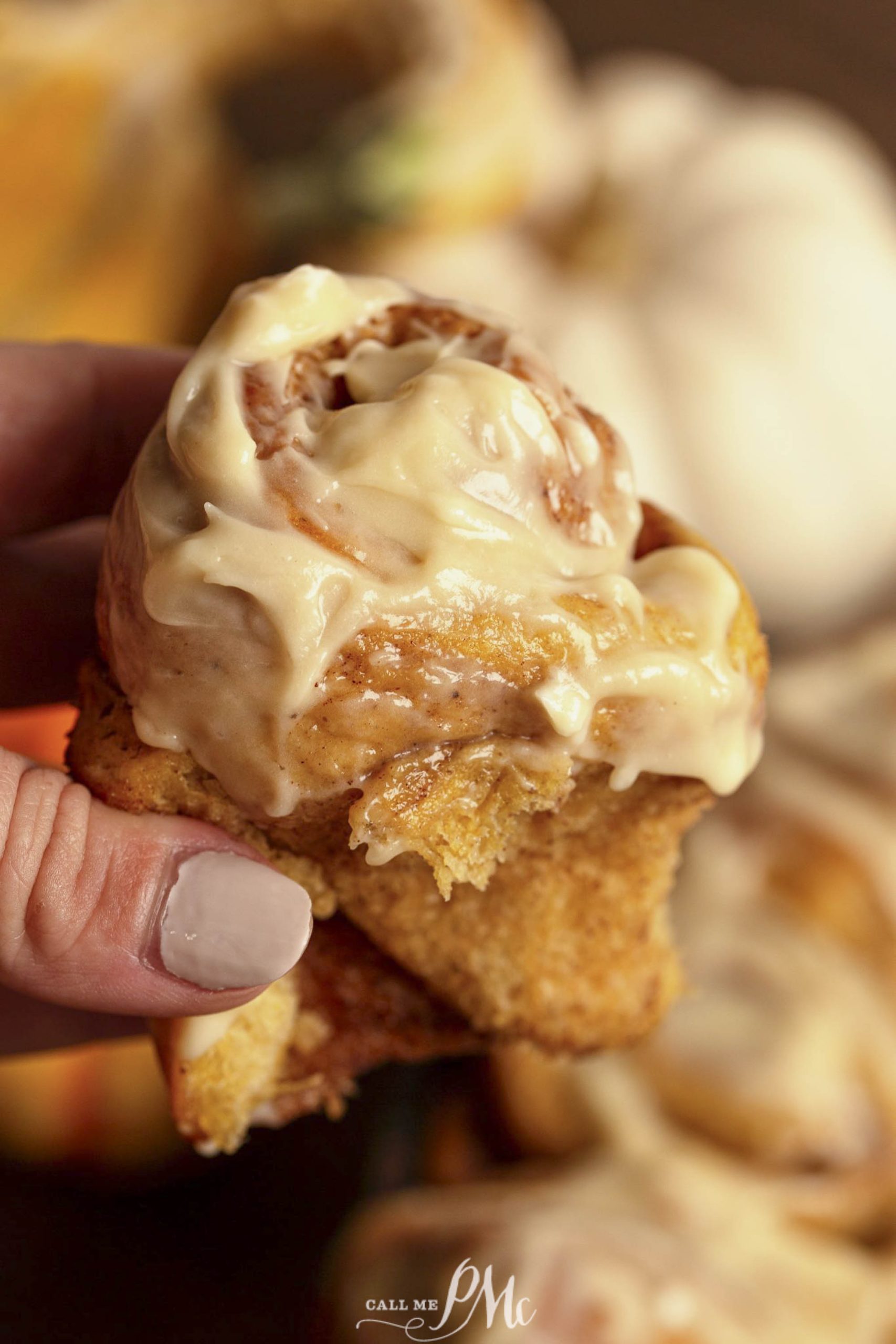 A close-up of a person's hand holding a frosted cinnamon roll topped with creamy icing.