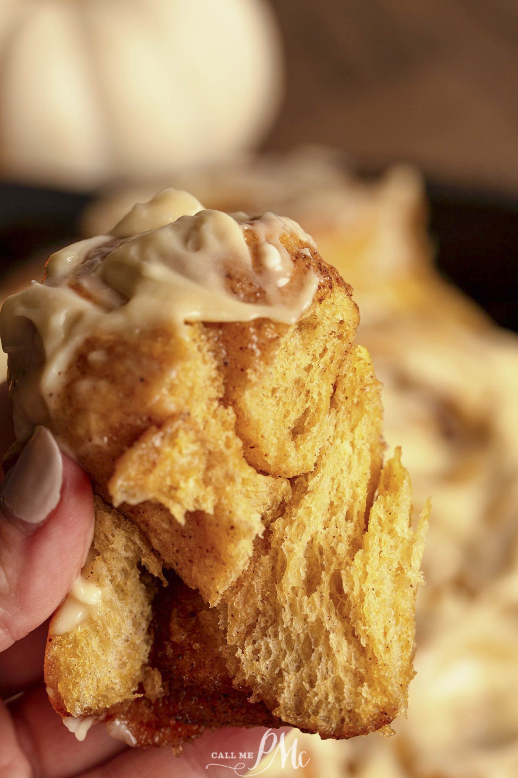 A close-up view of a hand holding a piece of cinnamon roll topped with glaze.