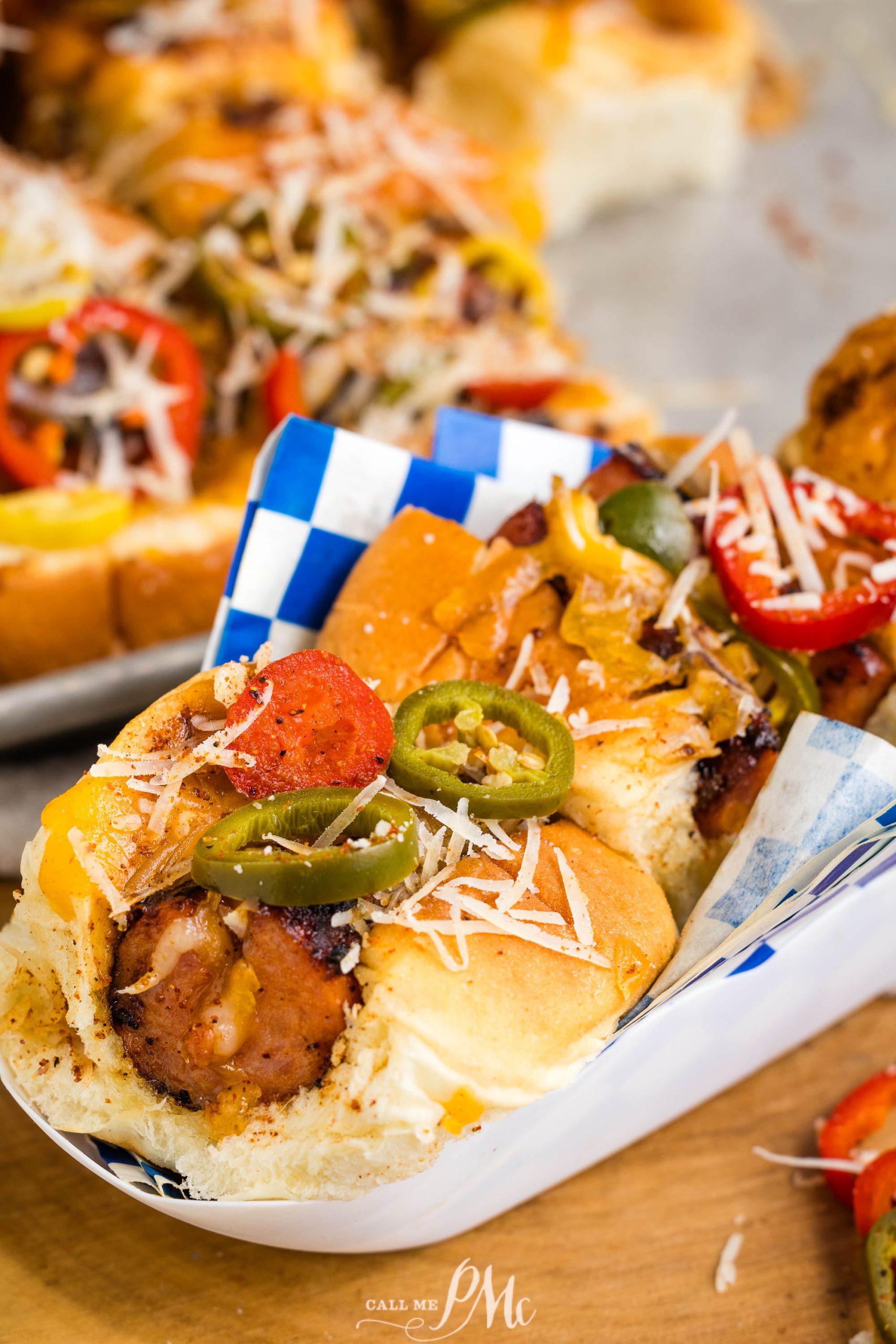 Close-up of a sandwich with grilled sausage, sliced bell peppers, jalapeños, and grated cheese in a blue and white checkered paper tray.