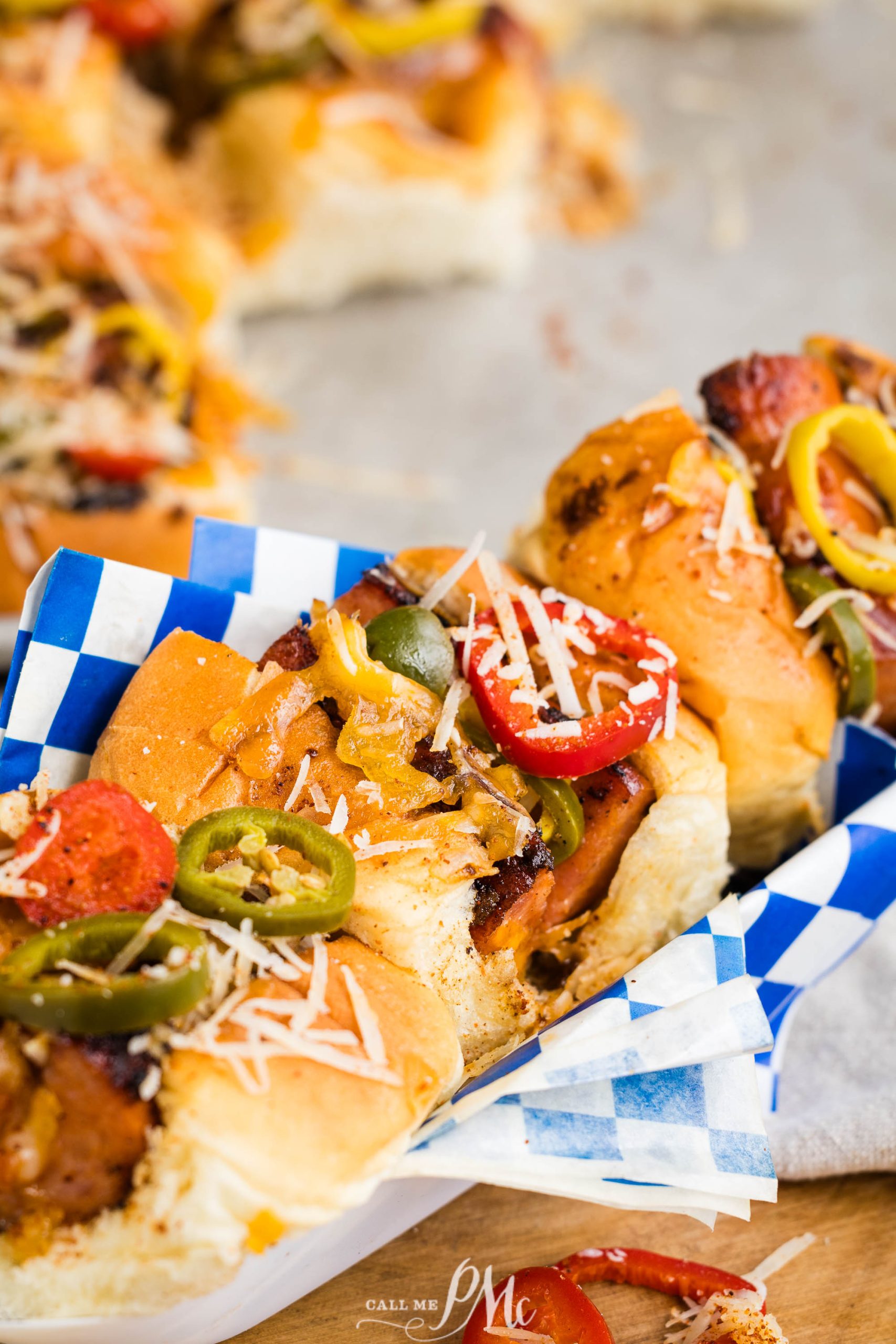 Close-up of sandwiches filled with sausages, jalapeños, pepperoni, cheese, and other toppings, wrapped in blue and white checkered paper.