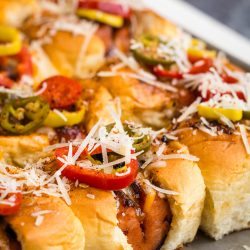 Close-up of baked sliders on a tray, topped with shredded cheese, sliced peppers, and spices.