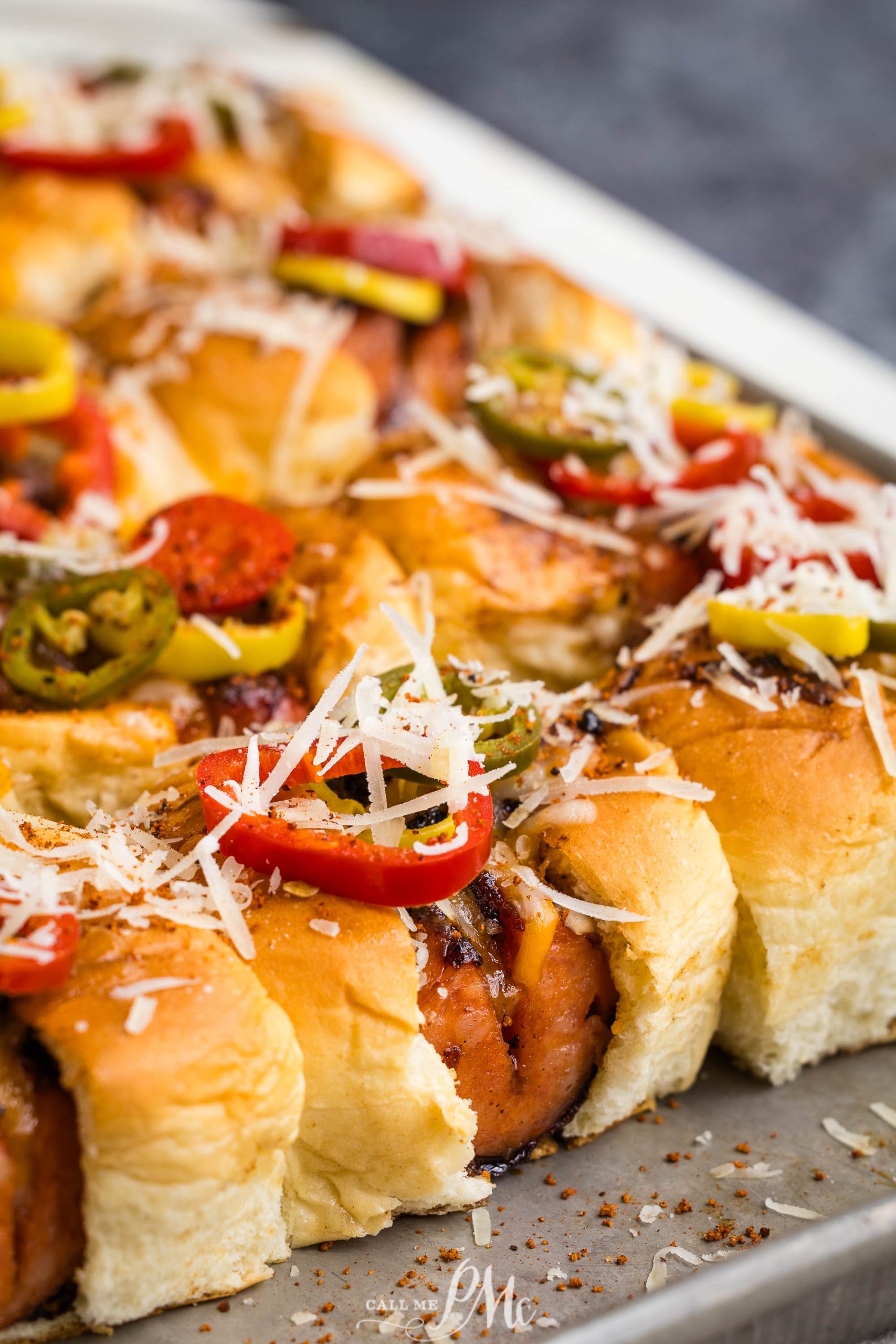 Close-up of baked sliders on a tray, topped with shredded cheese, sliced peppers, and spices.