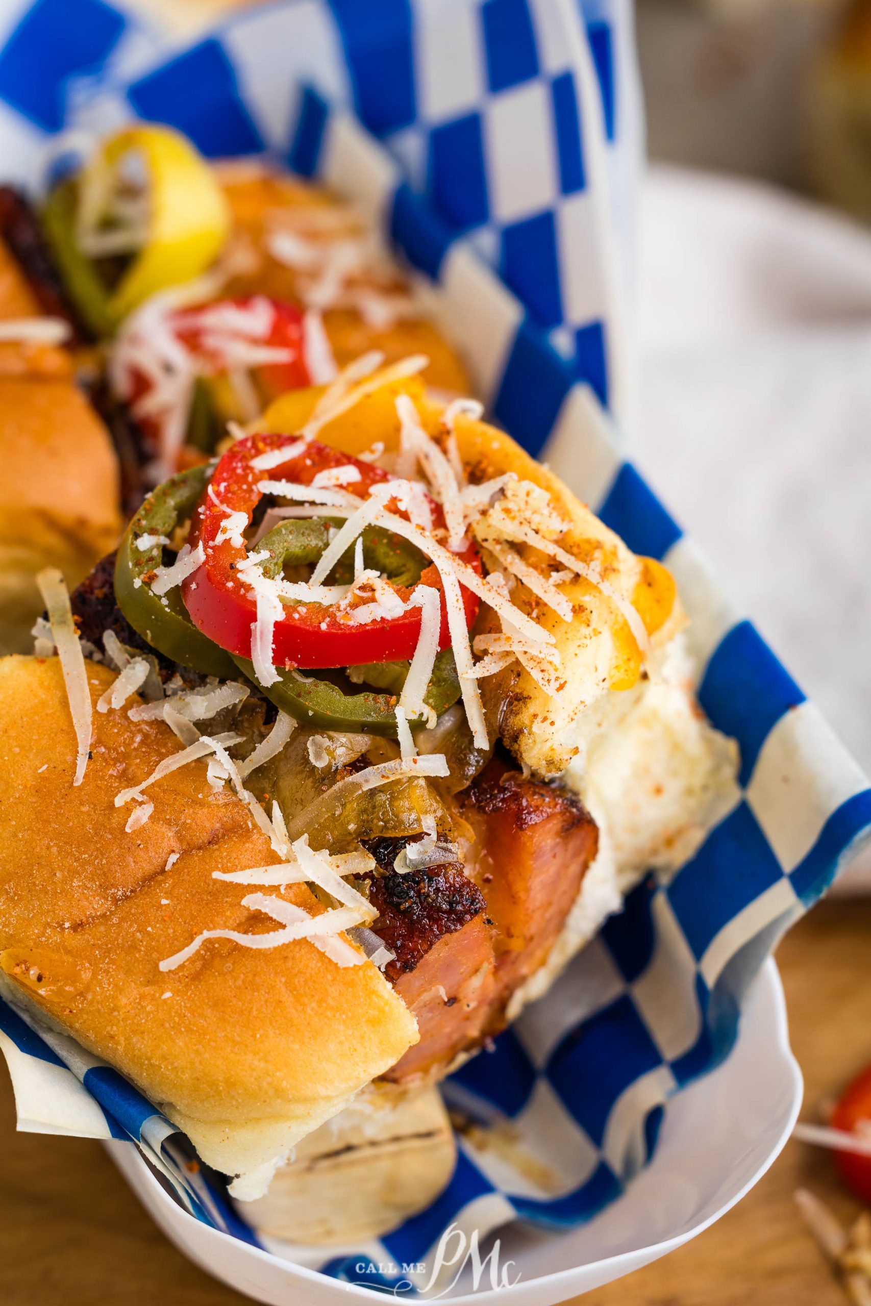 Close-up of a sandwich topped with grilled vegetables, grated cheese, and various seasonings, wrapped in blue and white checkered paper.