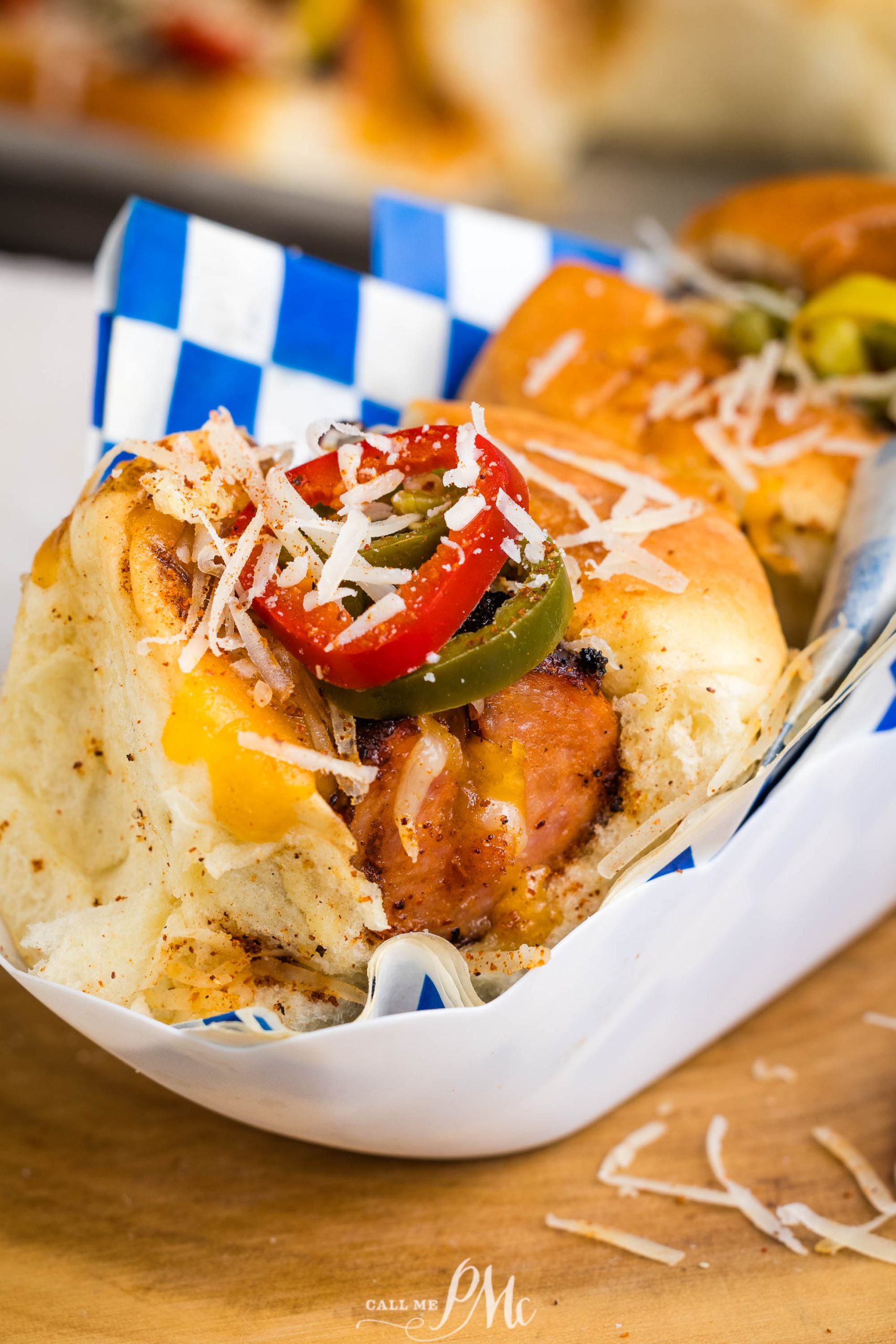 A close-up of sliders in a paper tray, topped with grilled sausage, melted cheese, sliced red and green peppers, and shredded cheese. The tray is lined with blue and white checkered paper.