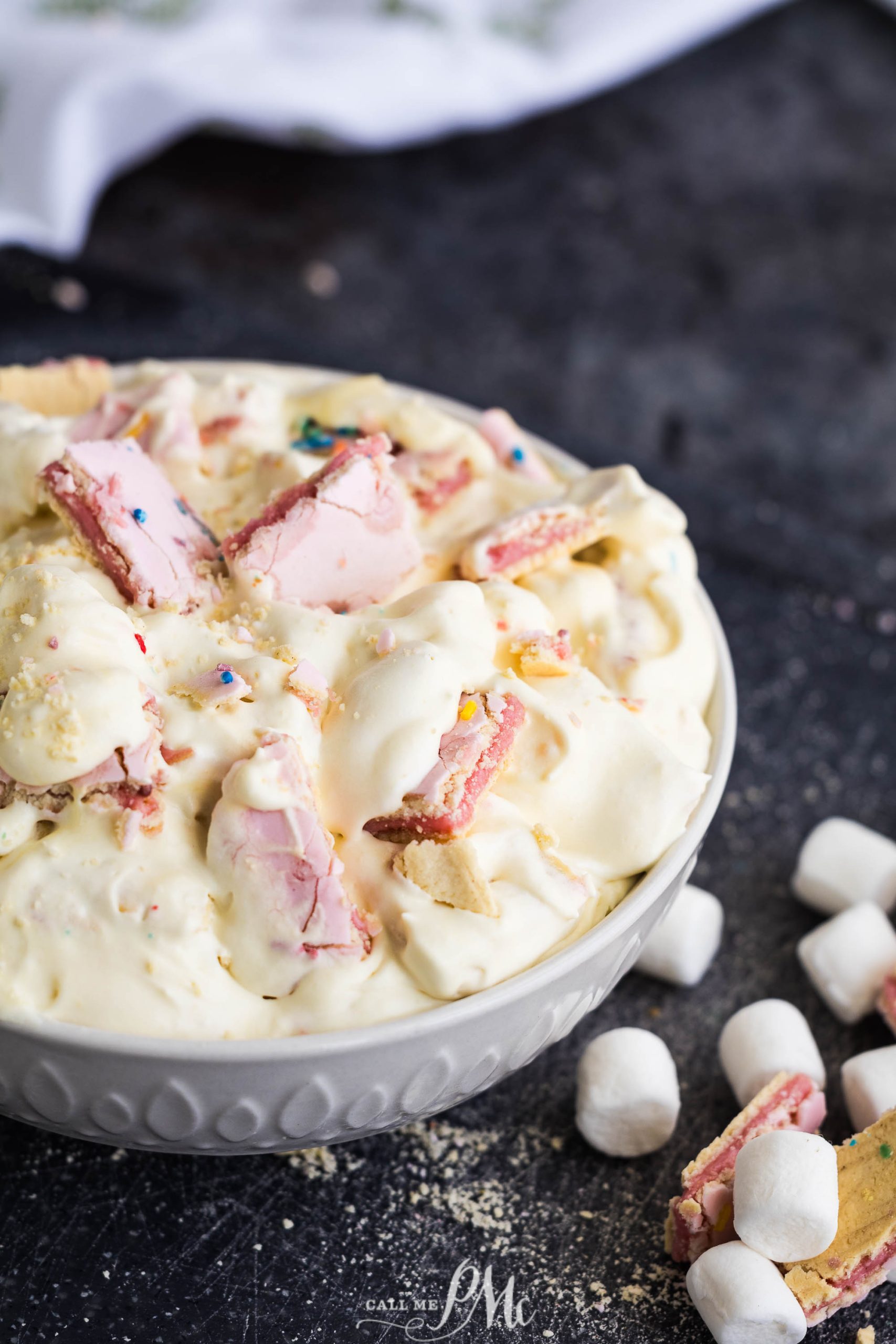 A bowl of creamy dessert topped with pieces of pink wafer cookies and mini marshmallows, with additional marshmallows scattered on the dark surface beside it.