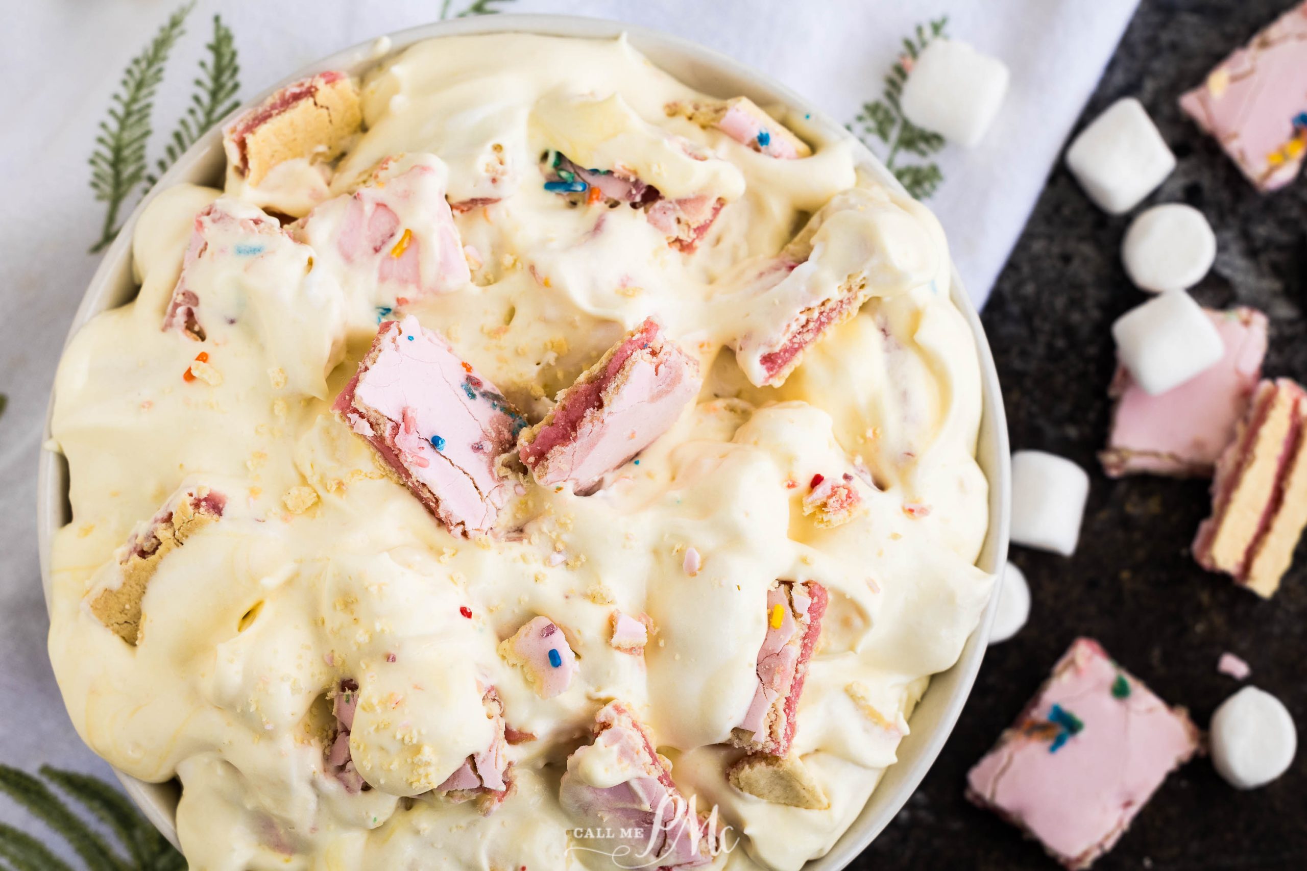 A bowl filled with creamy dessert topped with chunks of pink cookies, rainbow sprinkles, and small marshmallows on a white and black surface.