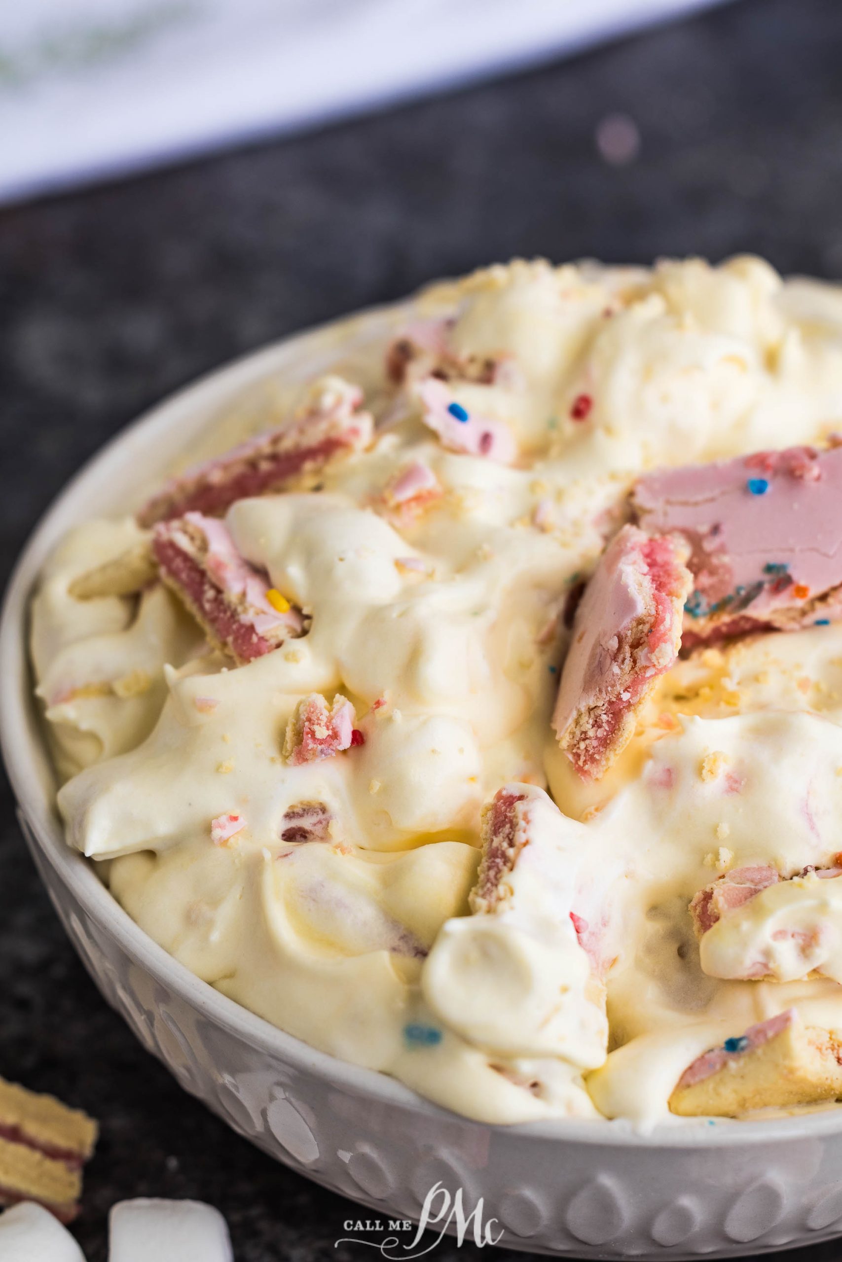 A bowl of creamy dessert with chunks of colorful candy and marshmallows on a dark surface.