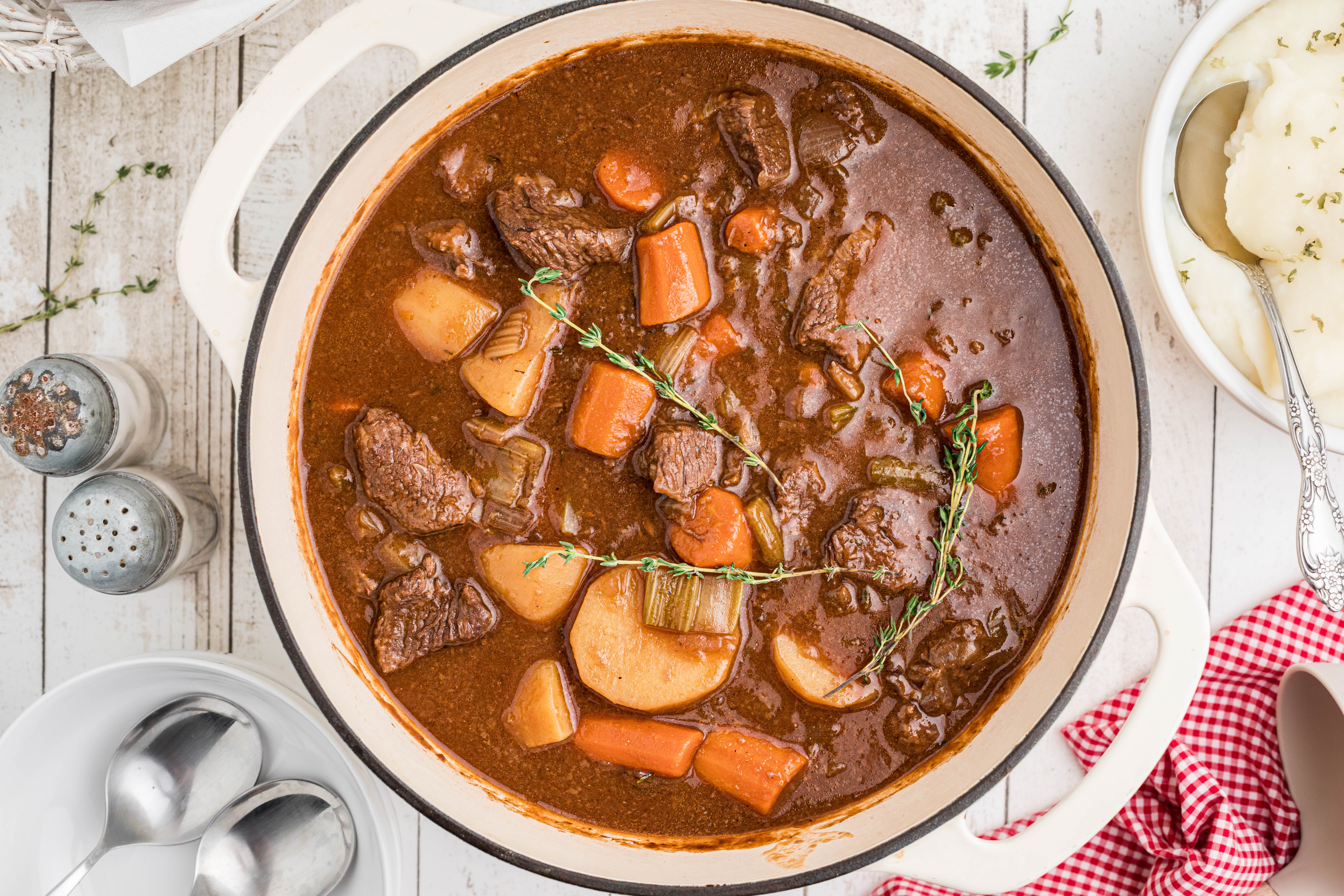 A bowl of beef stew with chunks of beef, carrots, and potatoes in a rich brown broth, garnished with fresh thyme. A metal spoon rests in the bowl.