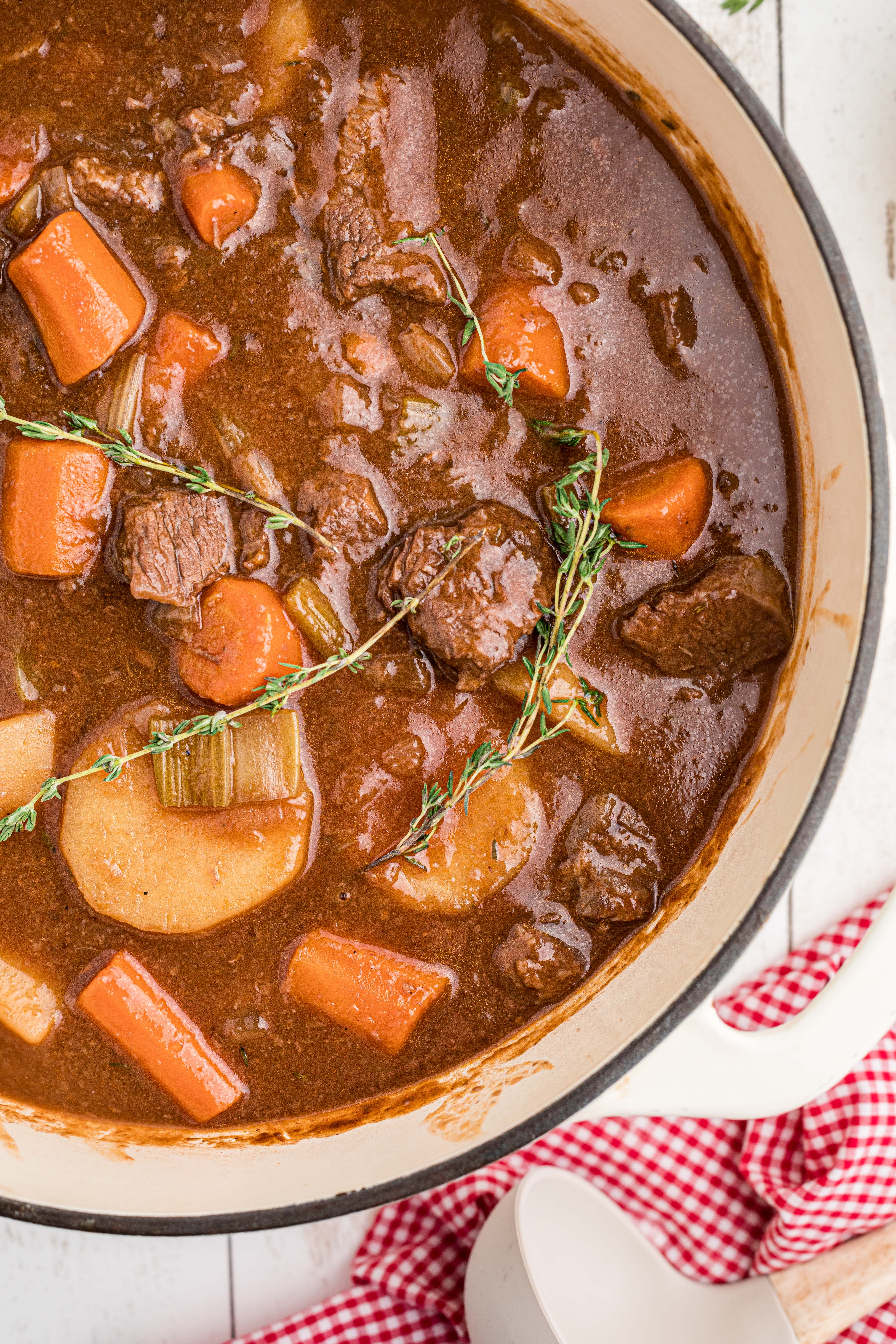 A bowl of beef stew with chunks of beef, carrots, and potatoes in a rich brown broth, garnished with fresh thyme. A metal spoon rests in the bowl.