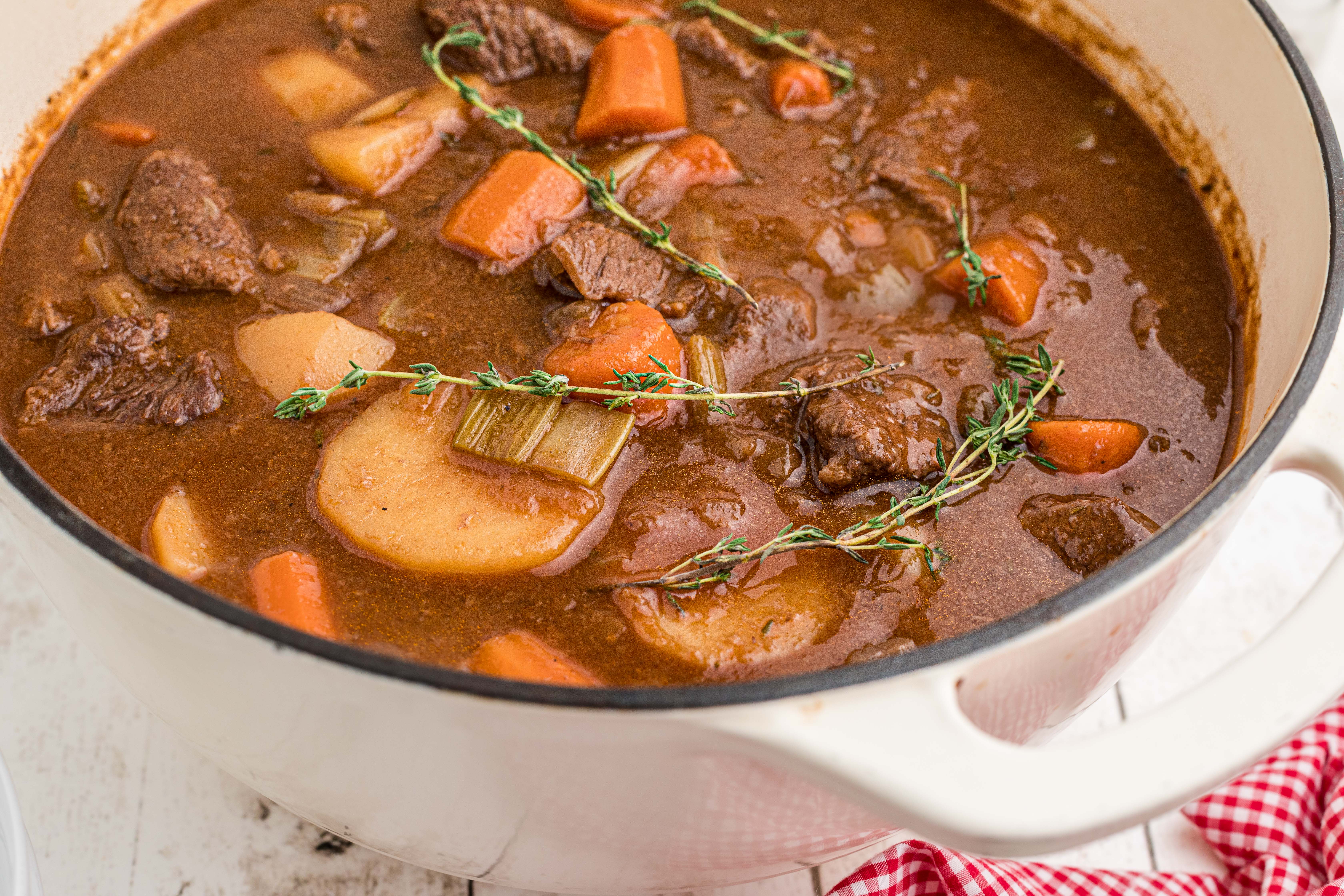 A bowl of beef stew with chunks of beef, carrots, and potatoes in a rich brown broth, garnished with fresh thyme. A metal spoon rests in the bowl.