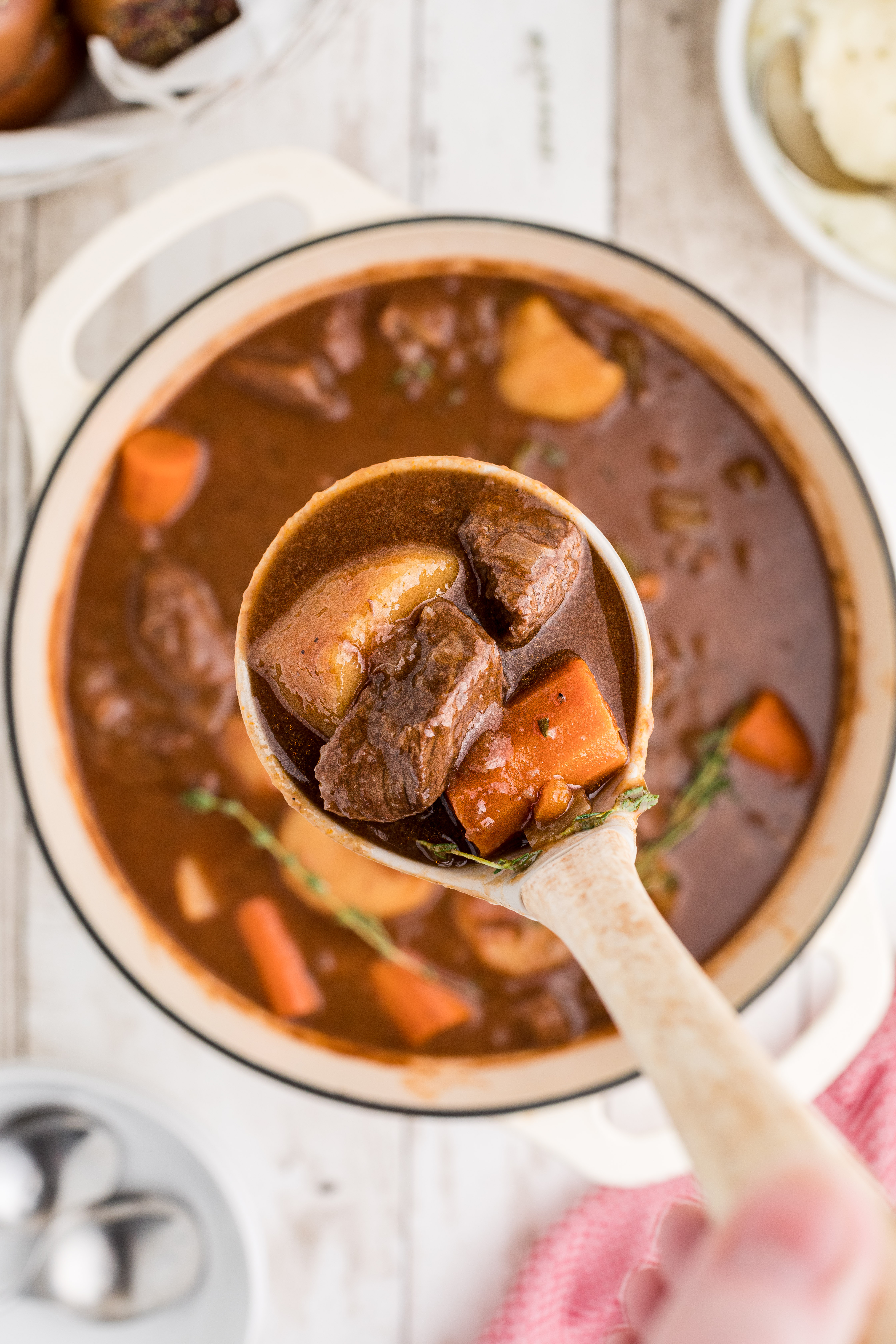 A bowl of beef stew with chunks of beef, carrots, and potatoes in a rich brown broth, garnished with fresh thyme. A metal spoon rests in the bowl.