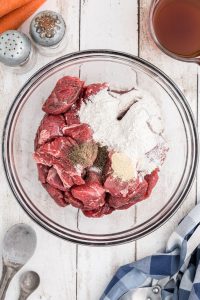 A glass bowl filled with raw beef chunks, flour, black pepper, and another seasoning on a white wooden surface. Nearby are salt and pepper shakers, a pitcher of broth, carrots, and utensils.