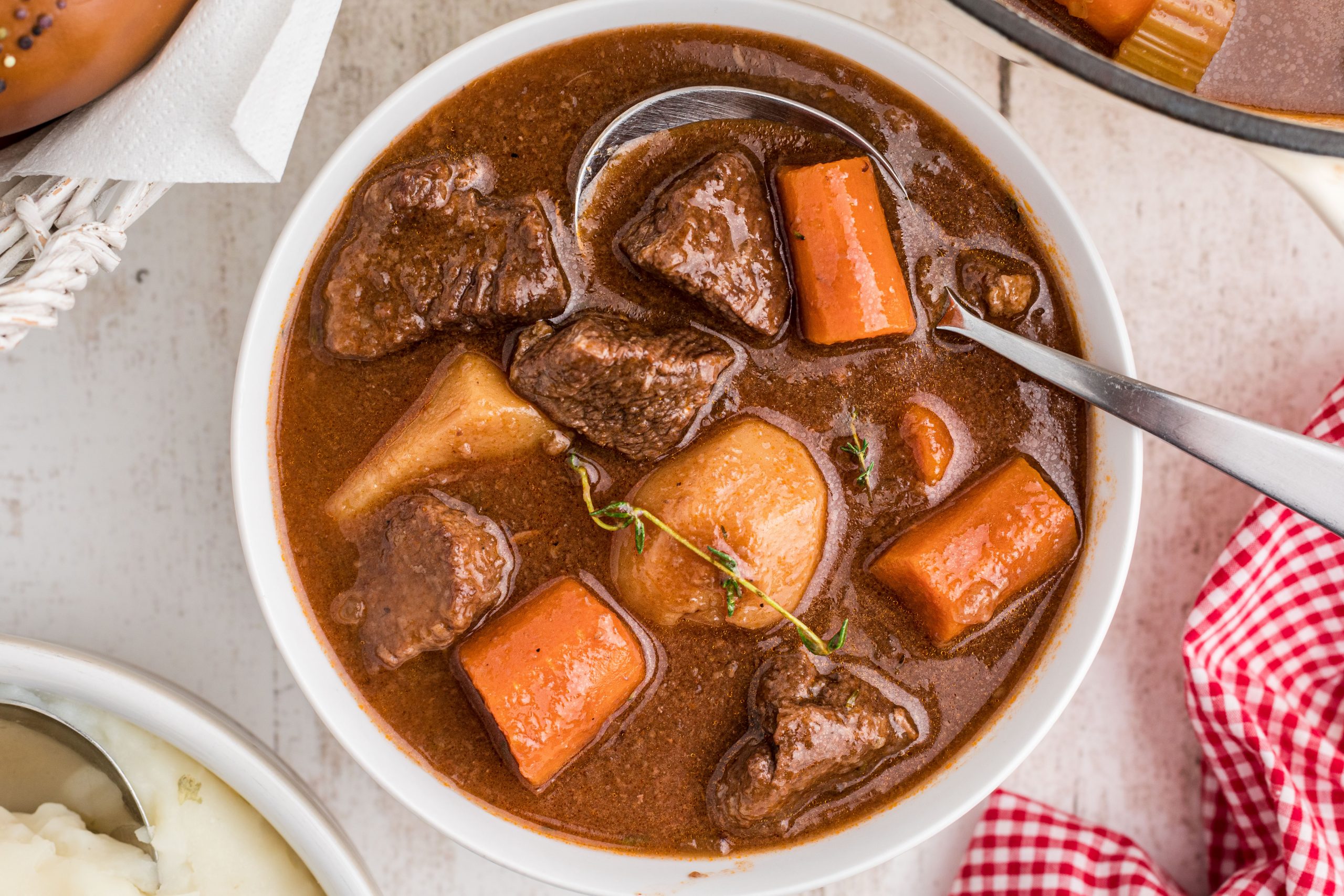 A bowl of beef stew with chunks of beef, carrots, and potatoes in a rich brown broth, garnished with fresh thyme. A metal spoon rests in the bowl.