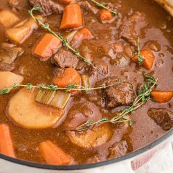 A white pot filled with traditional beef stew containing chunks of beef, carrots, potatoes, celery, and herbs in a rich brown sauce, placed on a red checkered cloth.