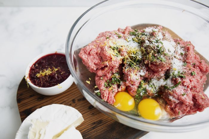 A bowl with ground meat, two eggs, herbs, and seasoning for mixing. A wooden board holds a small bowl of sauce and a round cheese.