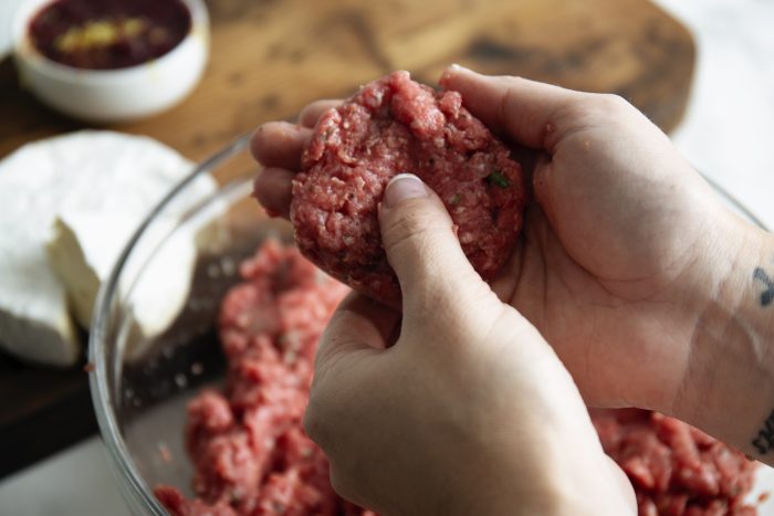 Close-up of hands shaping minced meat into a patty over a bowl, with a cutting board and white dish in the background.