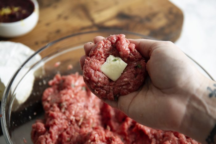 A hand holding seasoned ground meat with a piece of cheese placed in the center. A glass bowl with more ground meat is in the background.