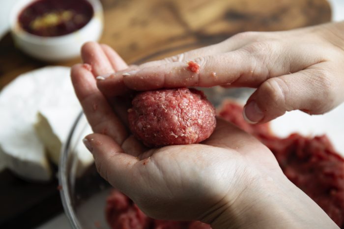 Hands shaping raw ground meat into a ball over a bowl, with ingredients blurred in the background.