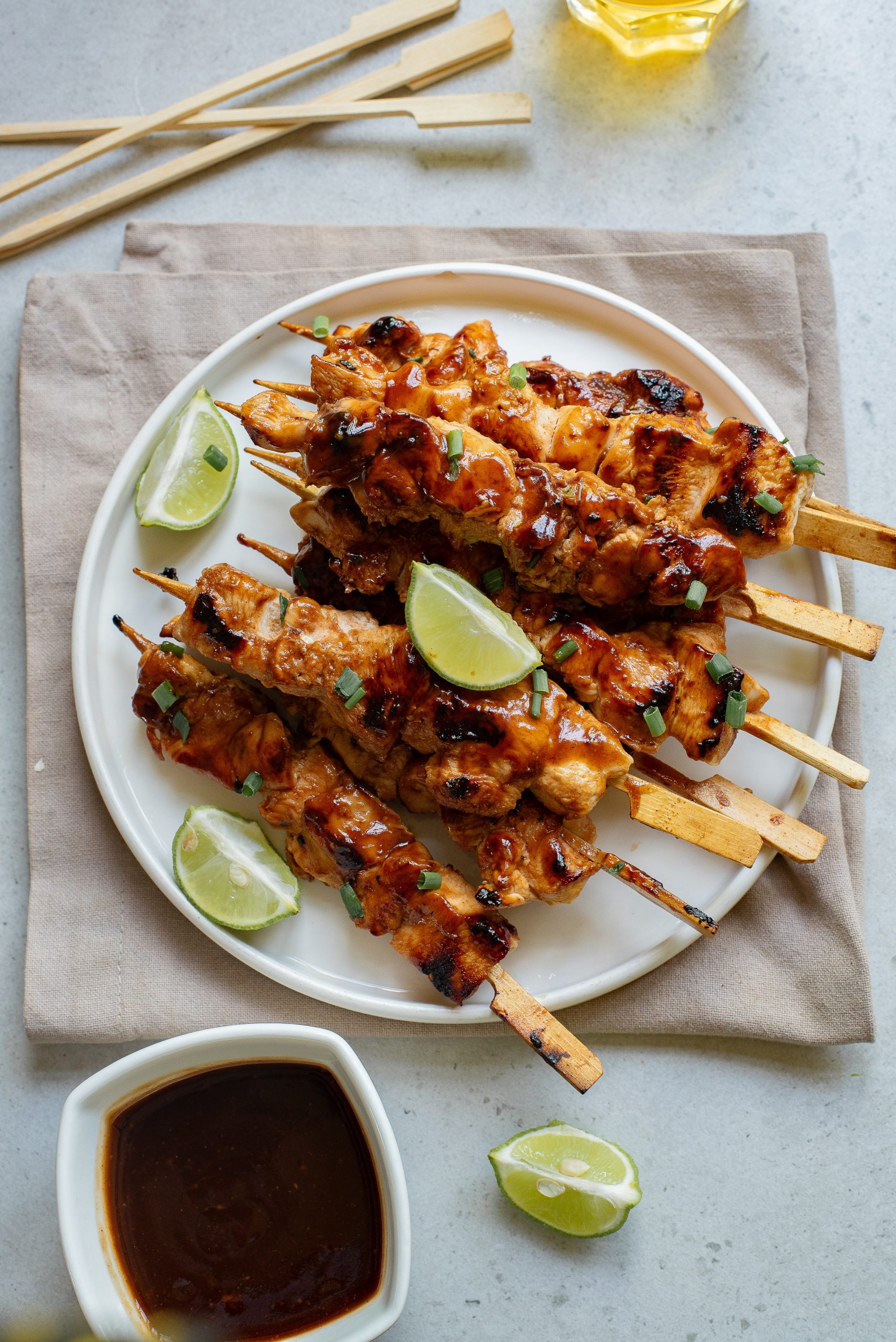 Grilled skewers of marinated chicken are served on a plate with lime wedges, accompanied by a small bowl of sauce and wooden sticks.