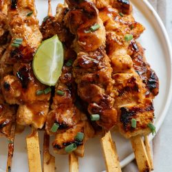 Grilled chicken skewers garnished with chopped green onions and a lime wedge, served on a white plate over a brown napkin.