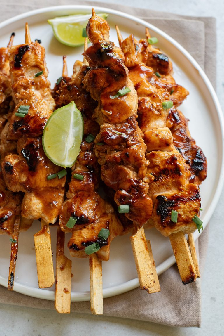 Grilled chicken skewers garnished with chopped green onions and a lime wedge, served on a white plate over a brown napkin.