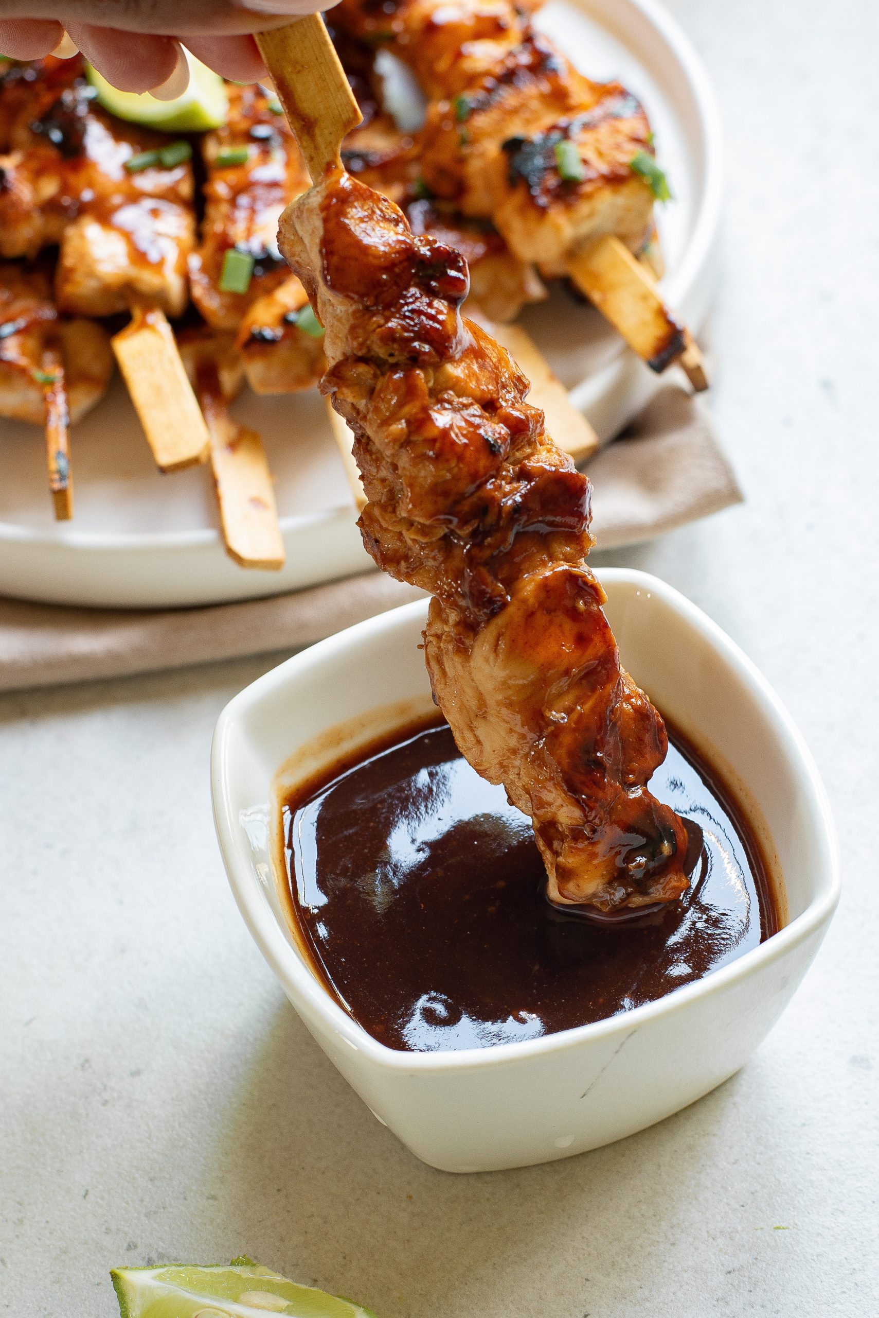 A chicken skewer is being dipped into a square bowl of dark sauce, with a plate of more skewers in the background.