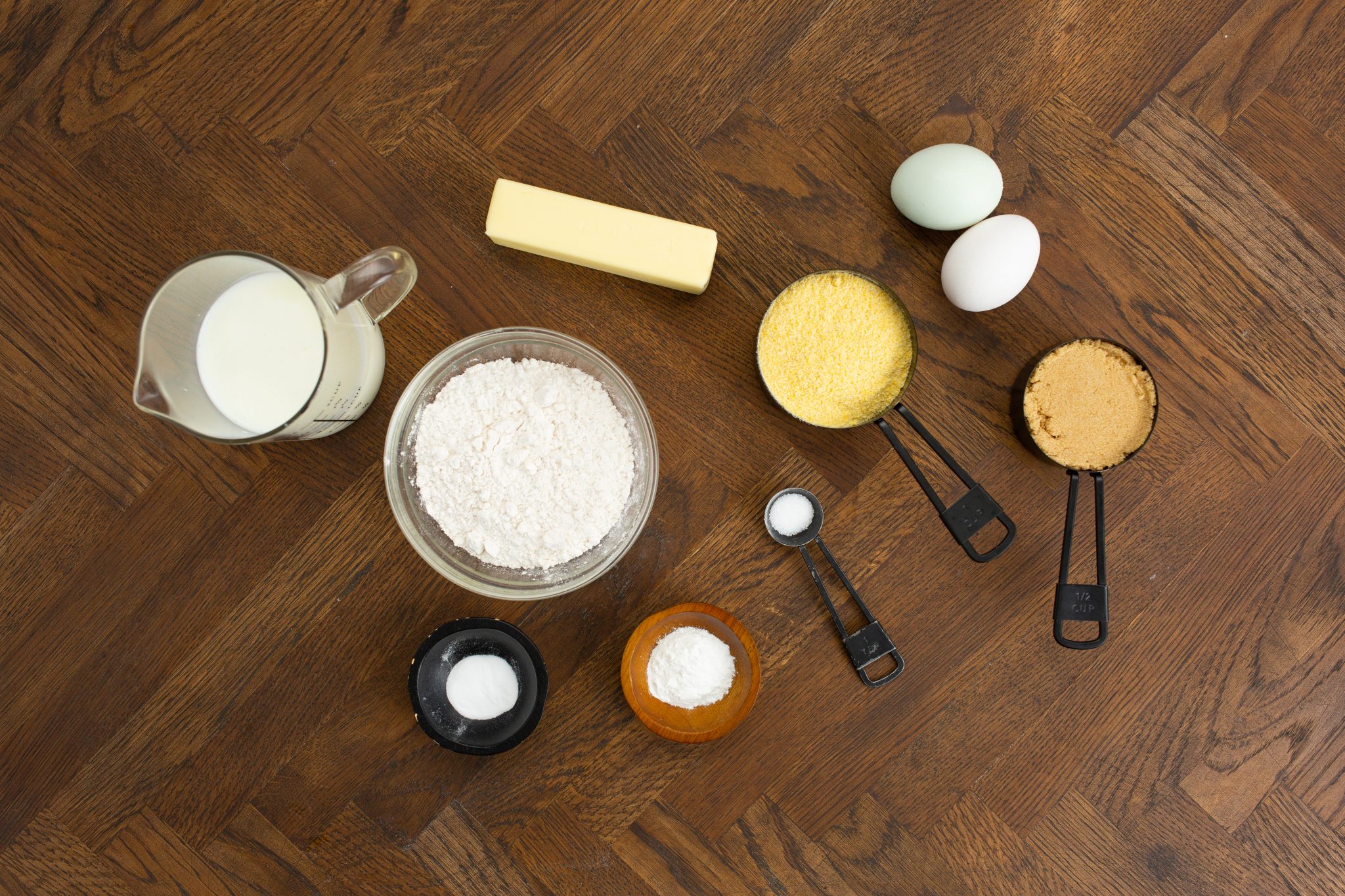 Baking ingredients on a wooden surface: milk, butter, eggs, flour, sugar, cornmeal, baking powder, and salt.