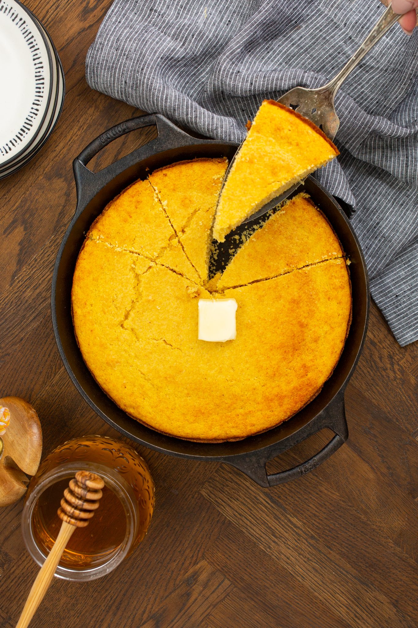 A cast iron skillet holds sliced Brown Sugar Cornbread with a pat of butter on top. One slice is being lifted out. Honey in a jar with a dipper is beside the skillet.