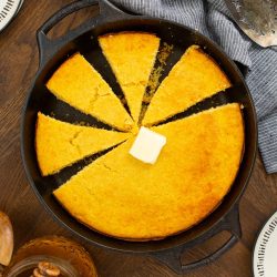 A sliced cornbread in a cast iron skillet with a pat of butter on top, a jar of honey nearby, and a striped cloth in the background.