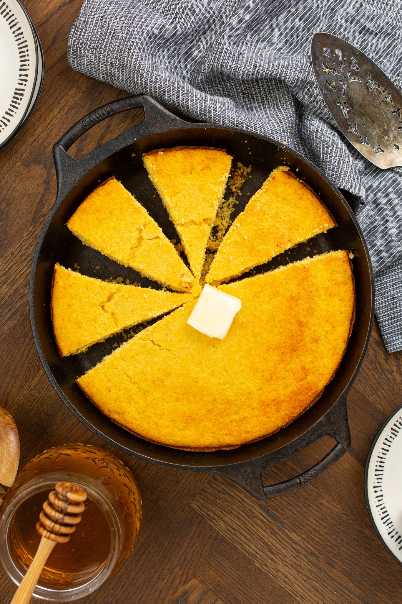 A sliced cornbread in a cast iron skillet with a pat of butter on top, a jar of honey nearby, and a striped cloth in the background.