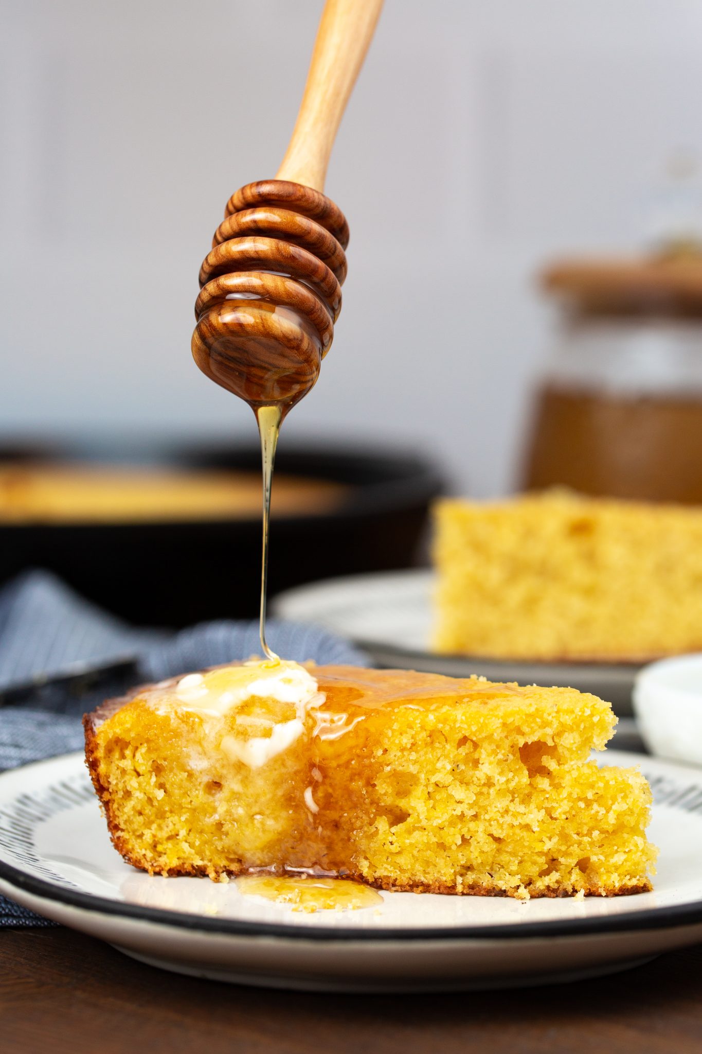 A slice of Brown Sugar Cornbread on a plate with honey drizzling from a wooden honey dipper above.