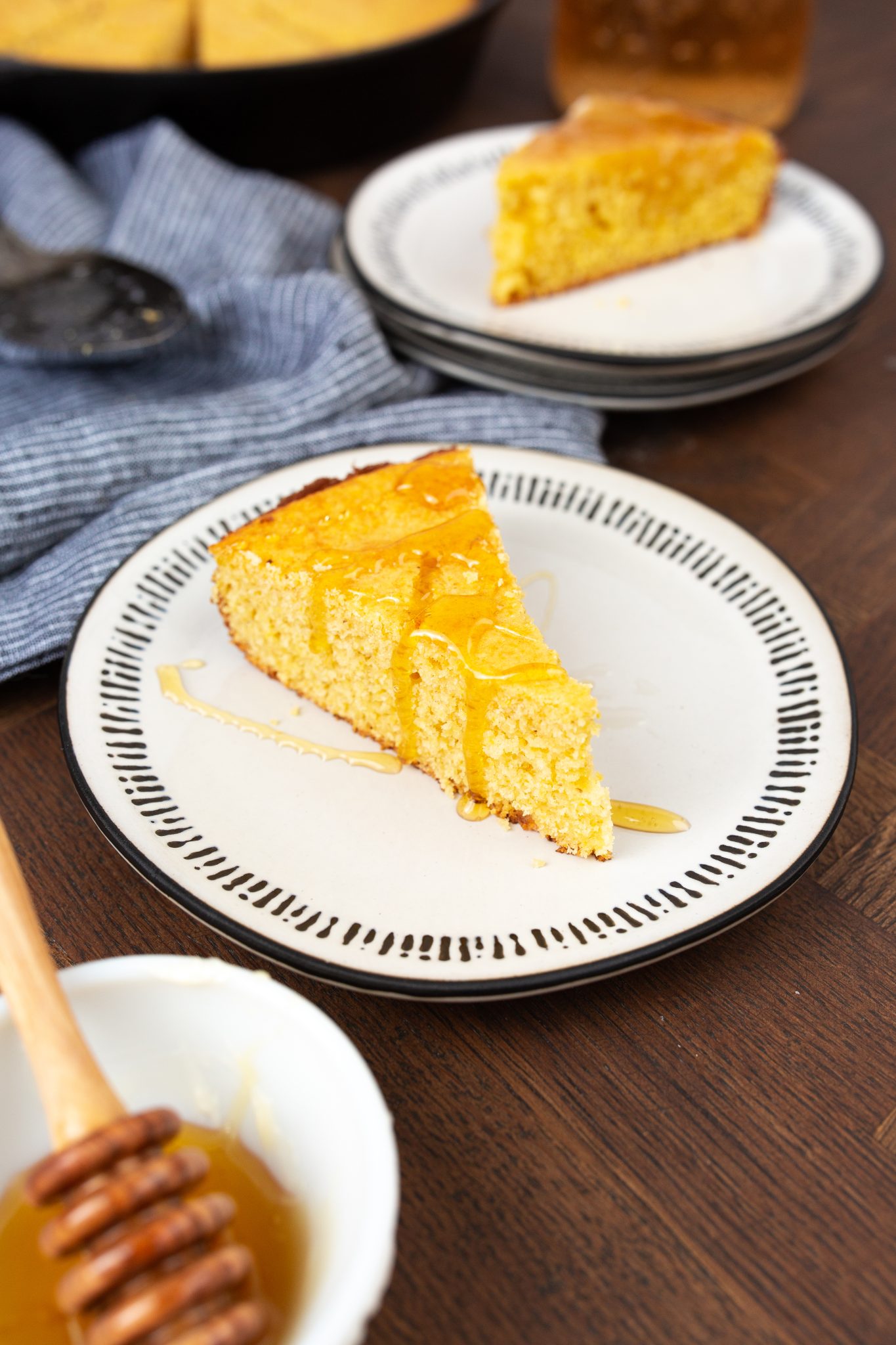 Slice of Brown Sugar Cornbread with honey on a plate, accompanied by a bowl with a honey dipper and stacked plates in the background.