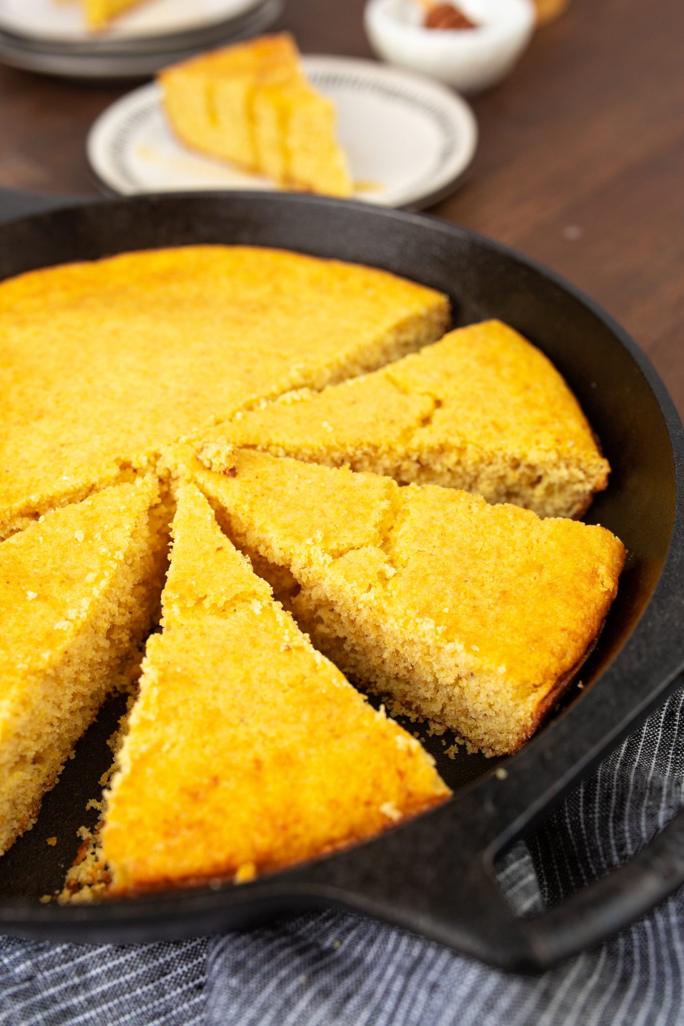 A cast iron skillet with sliced Brown Sugar Cornbread on a wooden table. Two plates with cornbread slices are in the background.