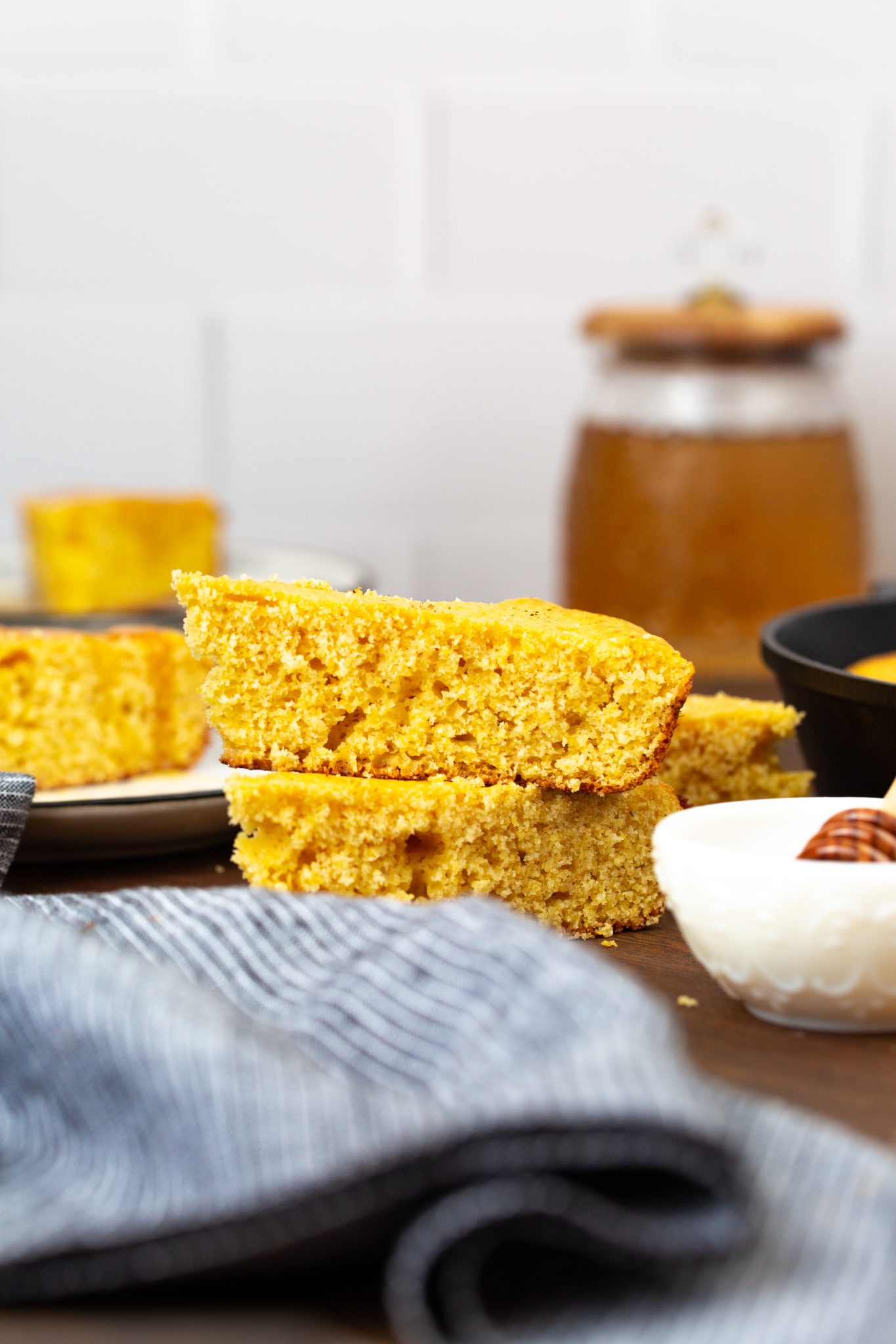 Slices of cornbread stacked on a plate with a jar of honey and a cloth napkin in the background.