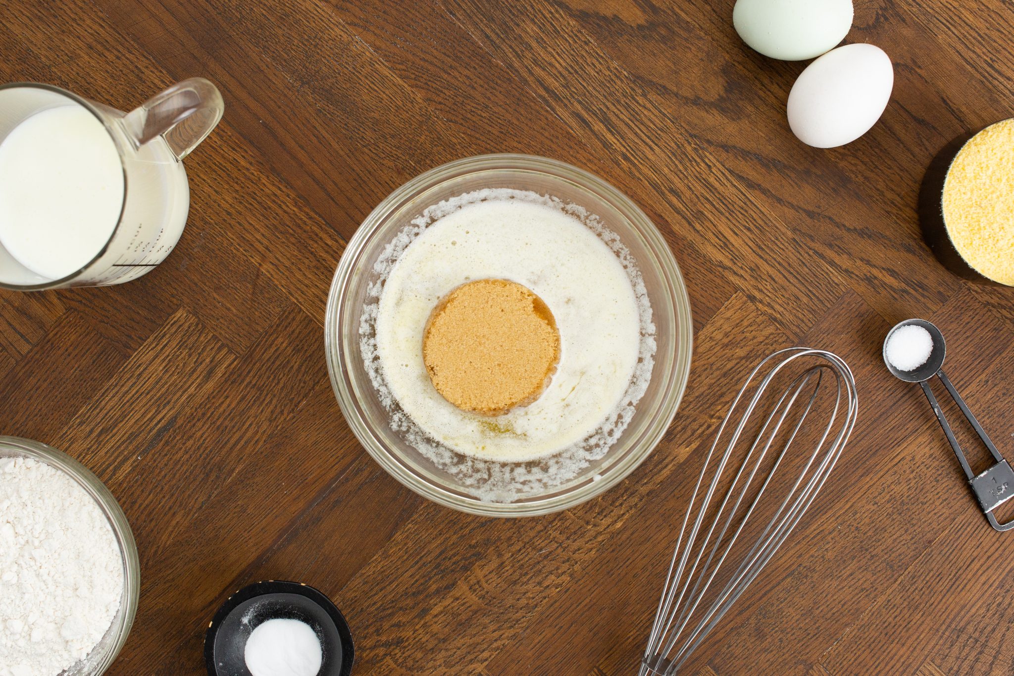 A mixing bowl with egg yolk and dry ingredients surrounded by milk, eggs, a whisk, cornmeal, flour, and baking powder on a wooden surface.