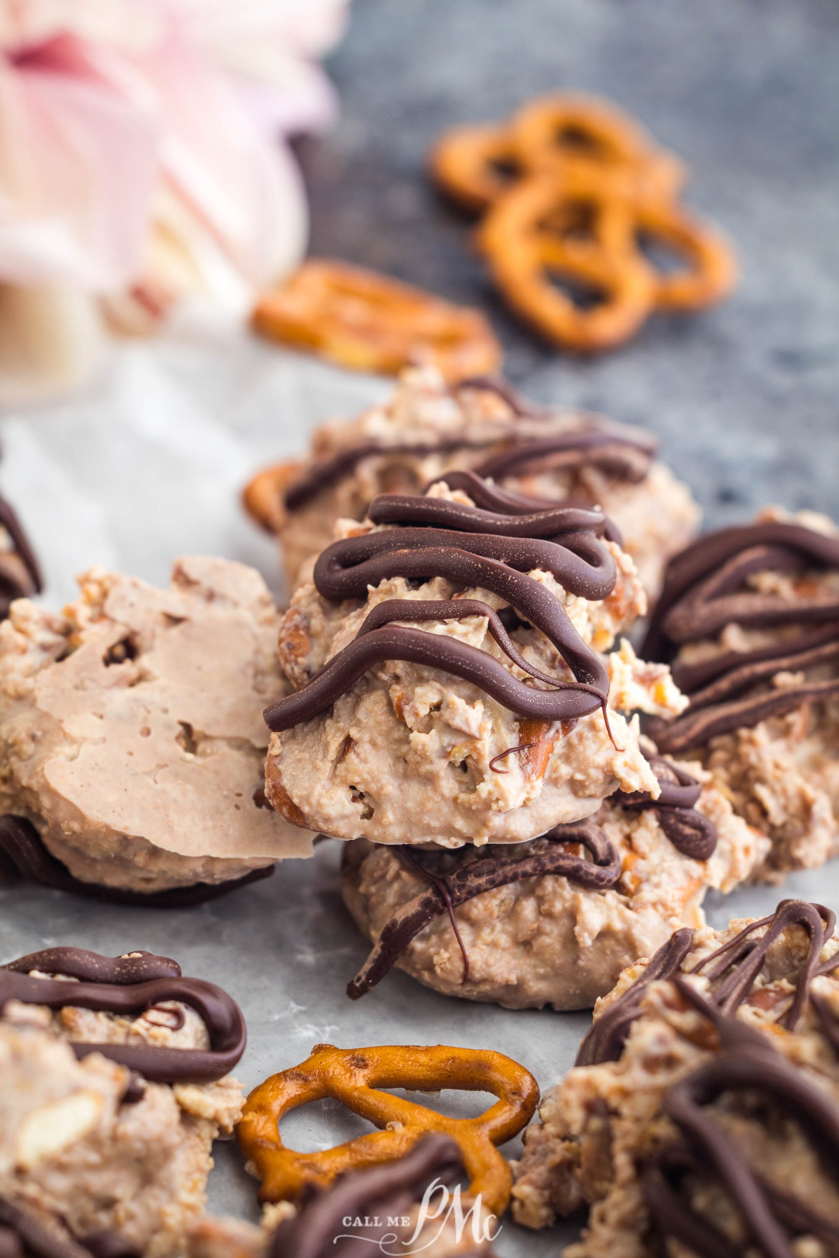 Close-up of cookies drizzled with chocolate, placed on parchment paper. Pretzels are scattered around the cookies.