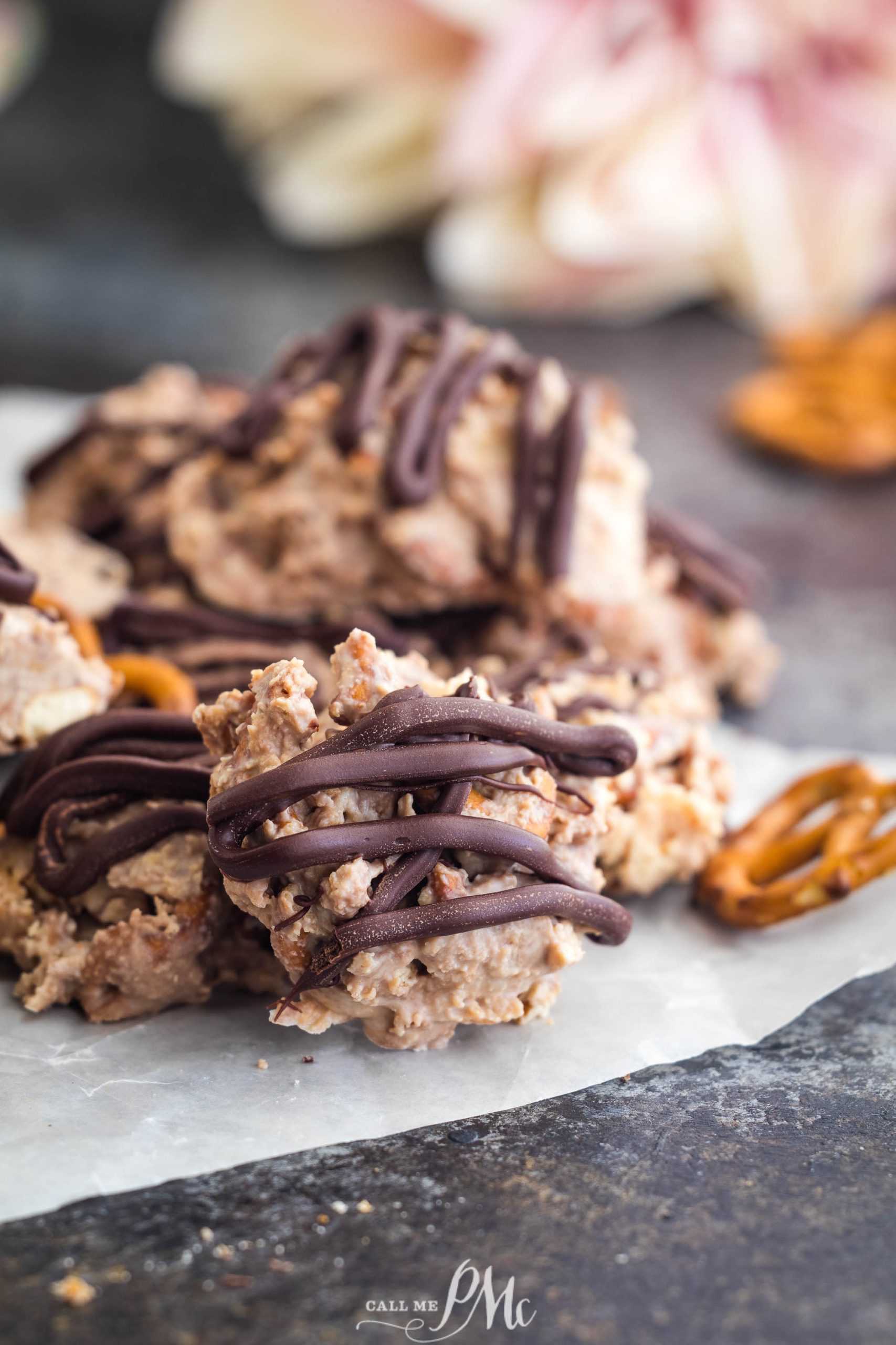 Chocolate-drizzled cookies piled on parchment paper with blurred background of floral decor and a pretzel.