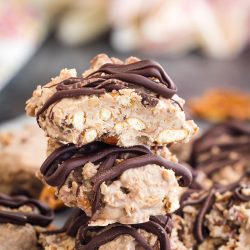 A close-up of stacked chocolate-drizzled peanut clusters with chunks of nuts visible. Blurred background with pink and white tones.