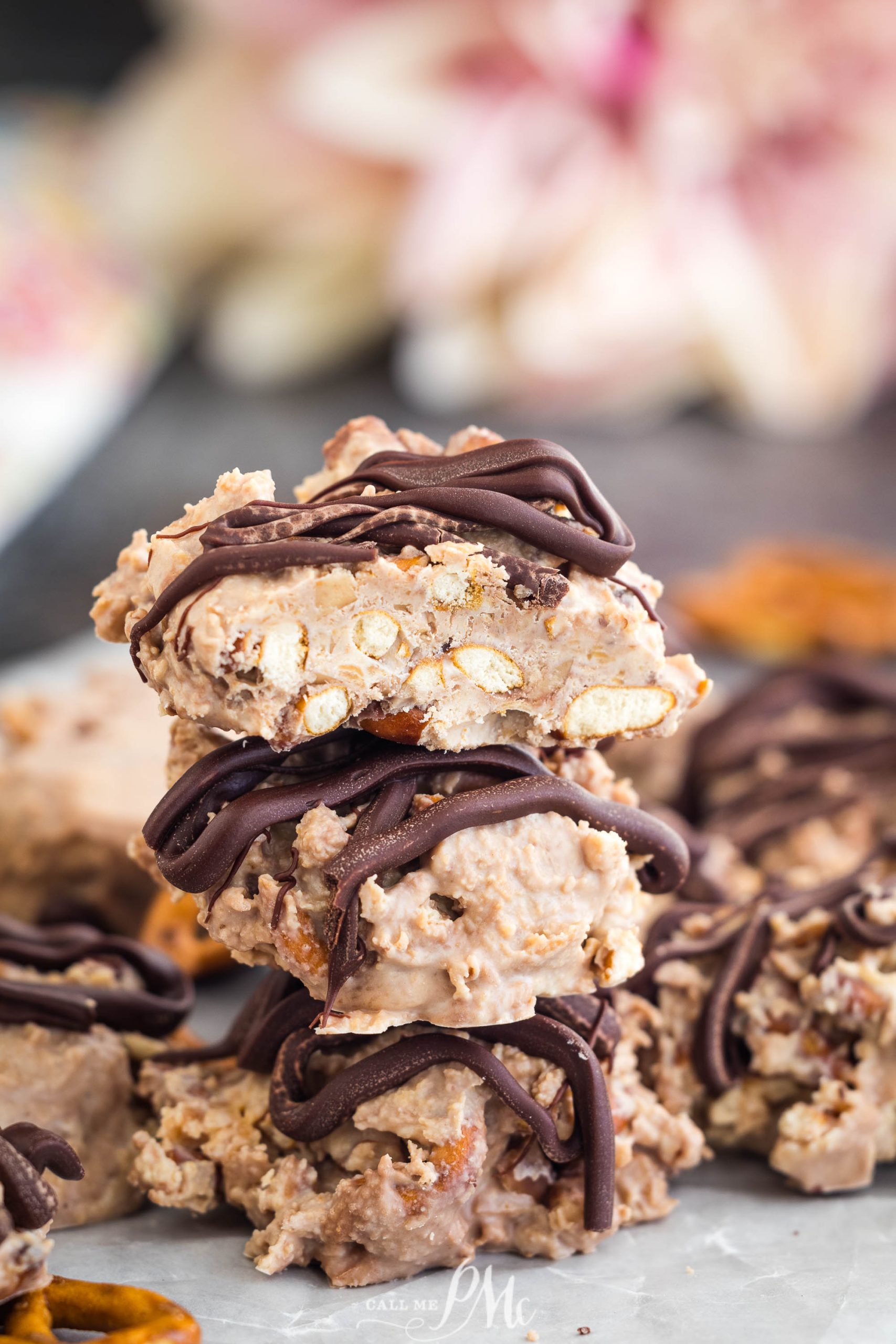 A close-up of stacked chocolate-drizzled peanut clusters with chunks of nuts visible. Blurred background with pink and white tones.