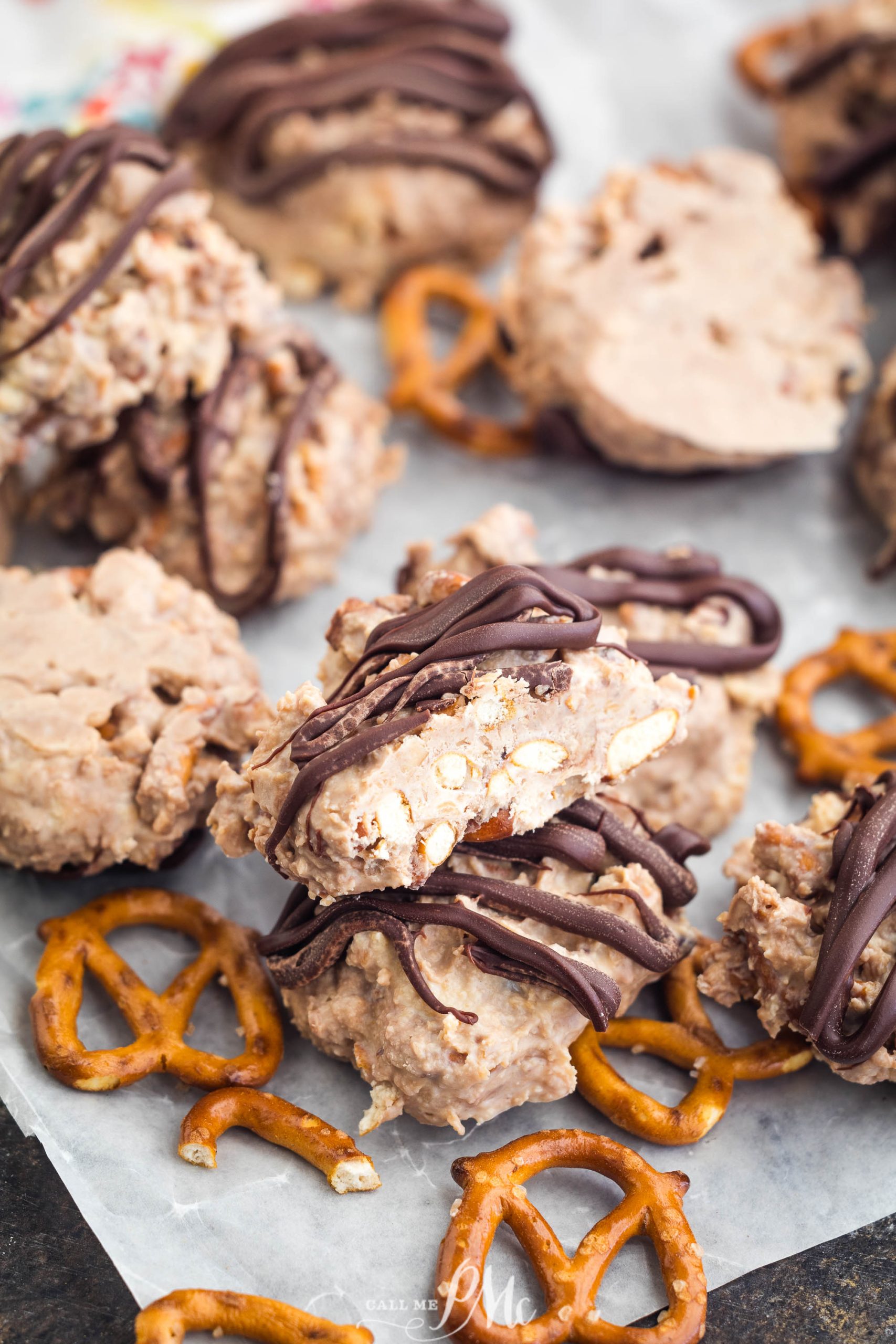 Cookies drizzled with chocolate, surrounded by pretzels, on parchment paper.