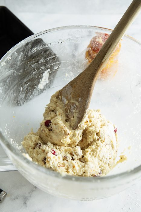 A wooden spoon in a bowl with dough mixture, containing visible chunks of cranberries and nuts, on a light countertop.