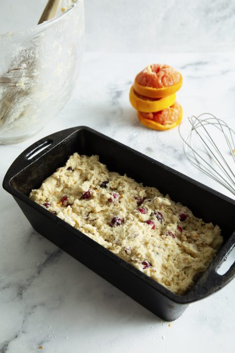 A loaf pan filled with unbaked cranberry bread batter sits on a countertop. Nearby are a mixing bowl, a whisk, and sliced oranges.