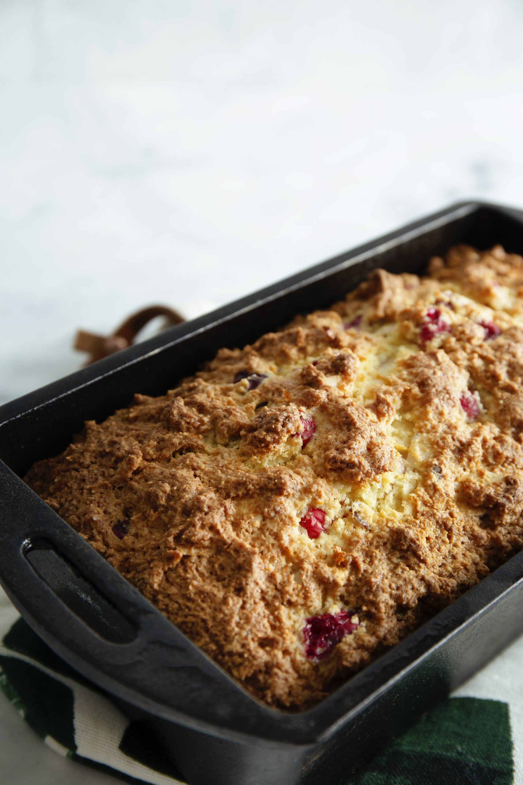 A loaf of Christmas Cranberry Bread with a golden-brown crust and visible berries, baked in a black rectangular pan, placed on a green and white cloth.