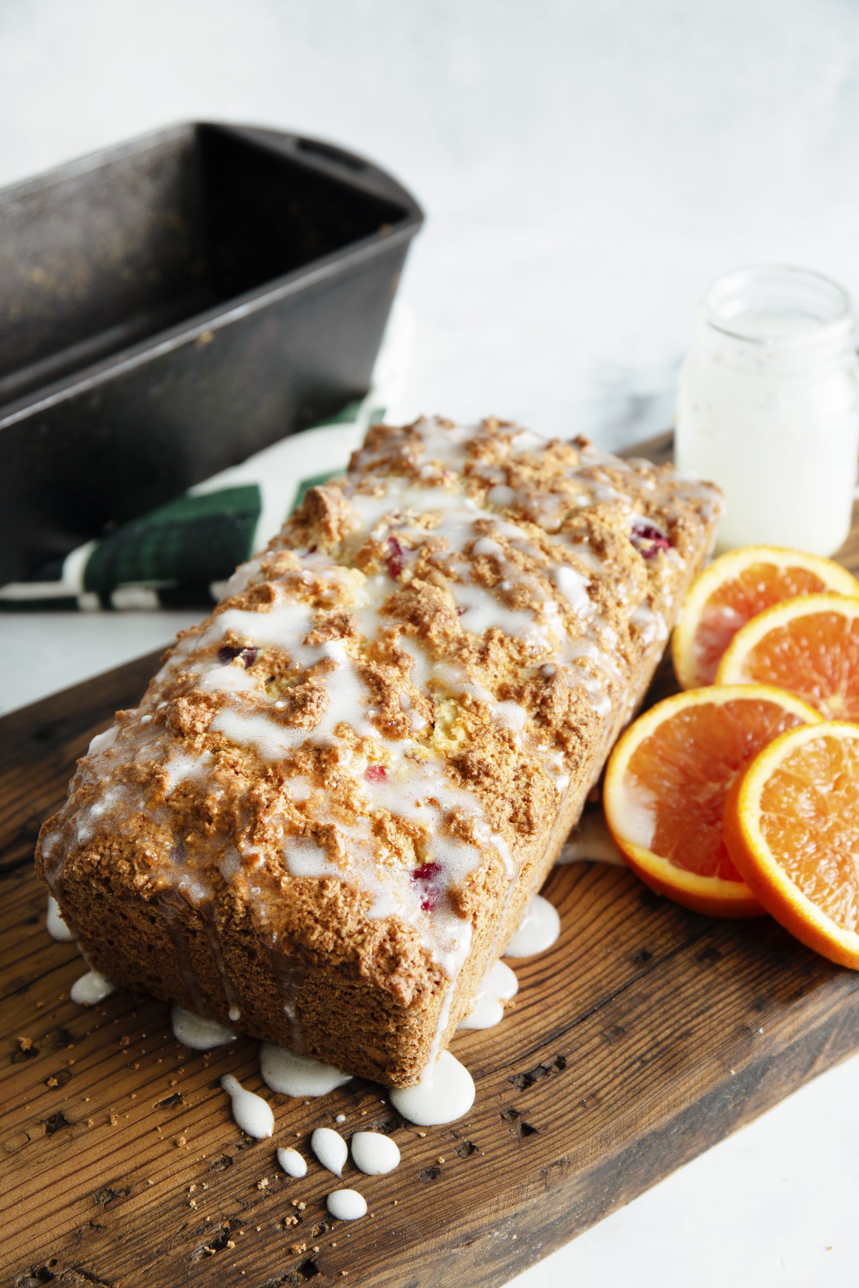 Loaf of Christmas Cranberry Bread with glaze on a wooden board, surrounded by orange slices. A baking pan and a jar are in the background.