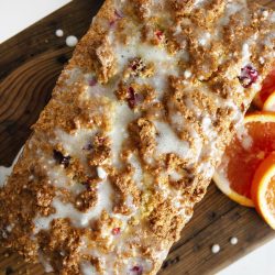 Loaf of citrus bread topped with glaze on a wooden cutting board, next to sliced oranges.