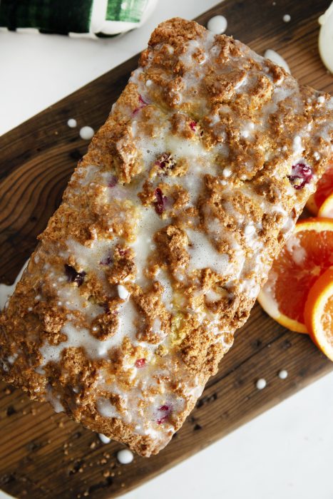 Loaf of Christmas Cranberry Bread topped with glaze on a wooden cutting board, next to sliced oranges.