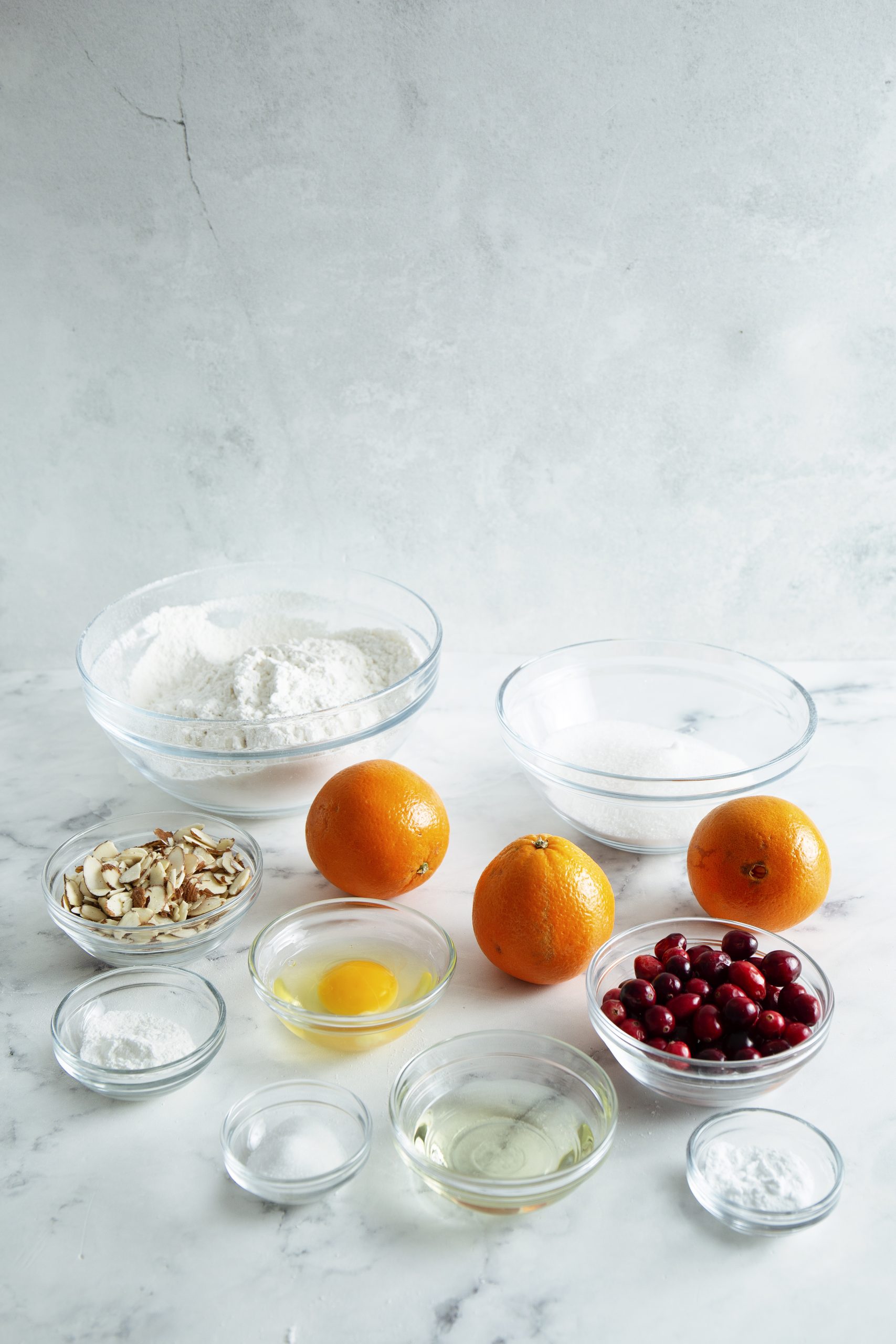 Various baking ingredients on a marble countertop, including flour, sugar, sliced almonds, two oranges, cranberries, an egg, oil, and small bowls of baking powder and salt.