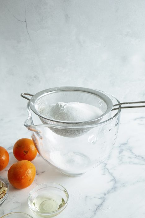 Sifter with flour over a glass bowl, surrounded by oranges and small bowls of ingredients on a marble surface.