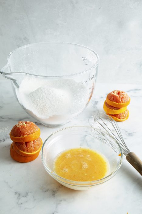 Bowl of flour, a whisk, squeezed oranges, and a bowl of beaten eggs on a marble countertop.